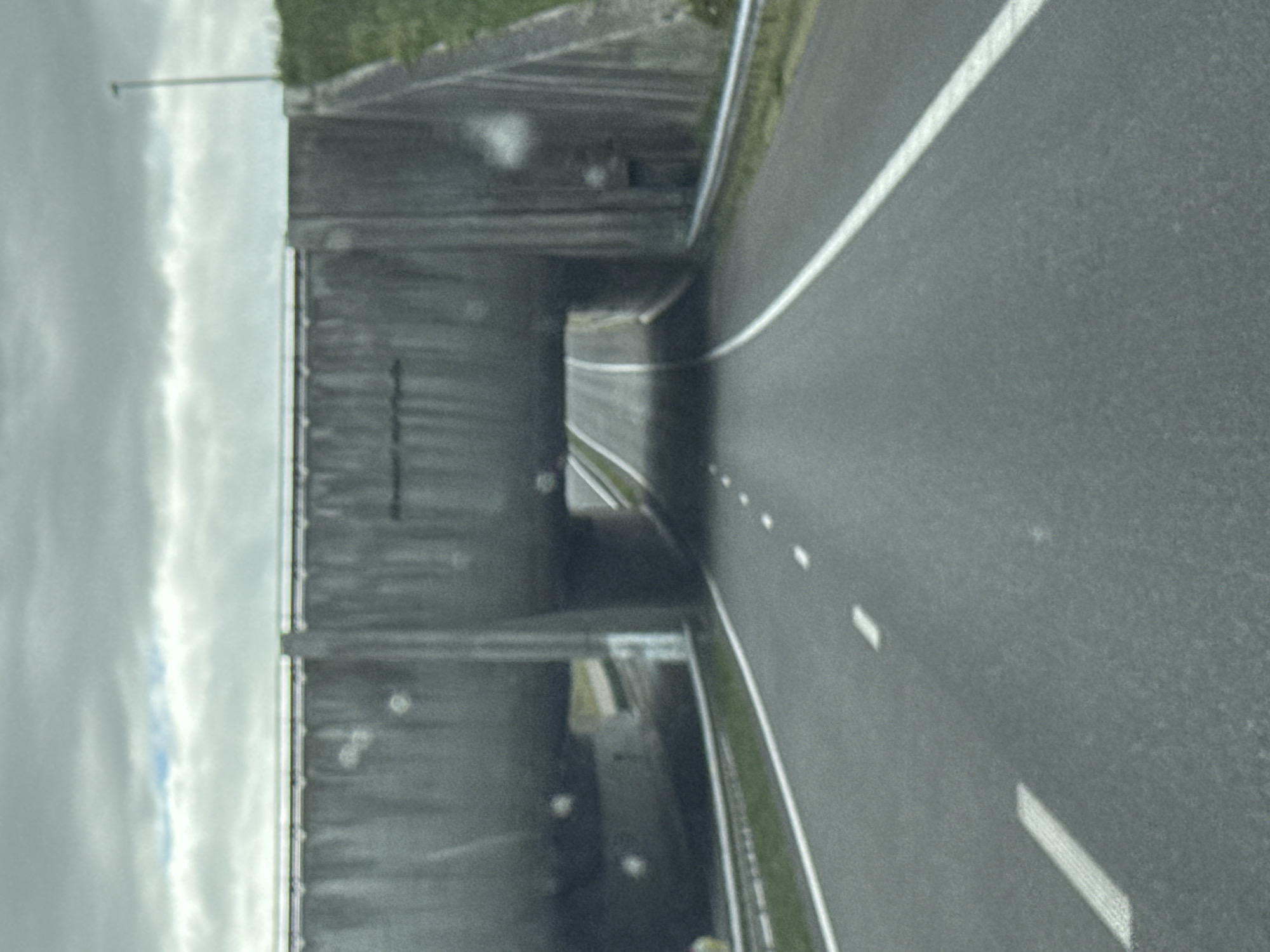 The image shows a highway passing under a concrete wildlife overpass in Grou, Netherlands. The overpass is a large, grey structure with sloping sides and a flat top, covered in grass on the right side of the image. The highway itself is asphalt, with white lane markings. The sky is overcast, with some patches of blue visible through the clouds. There are no people or vehicles visible in the image. The scene is captured from the perspective of someone driving on the highway.