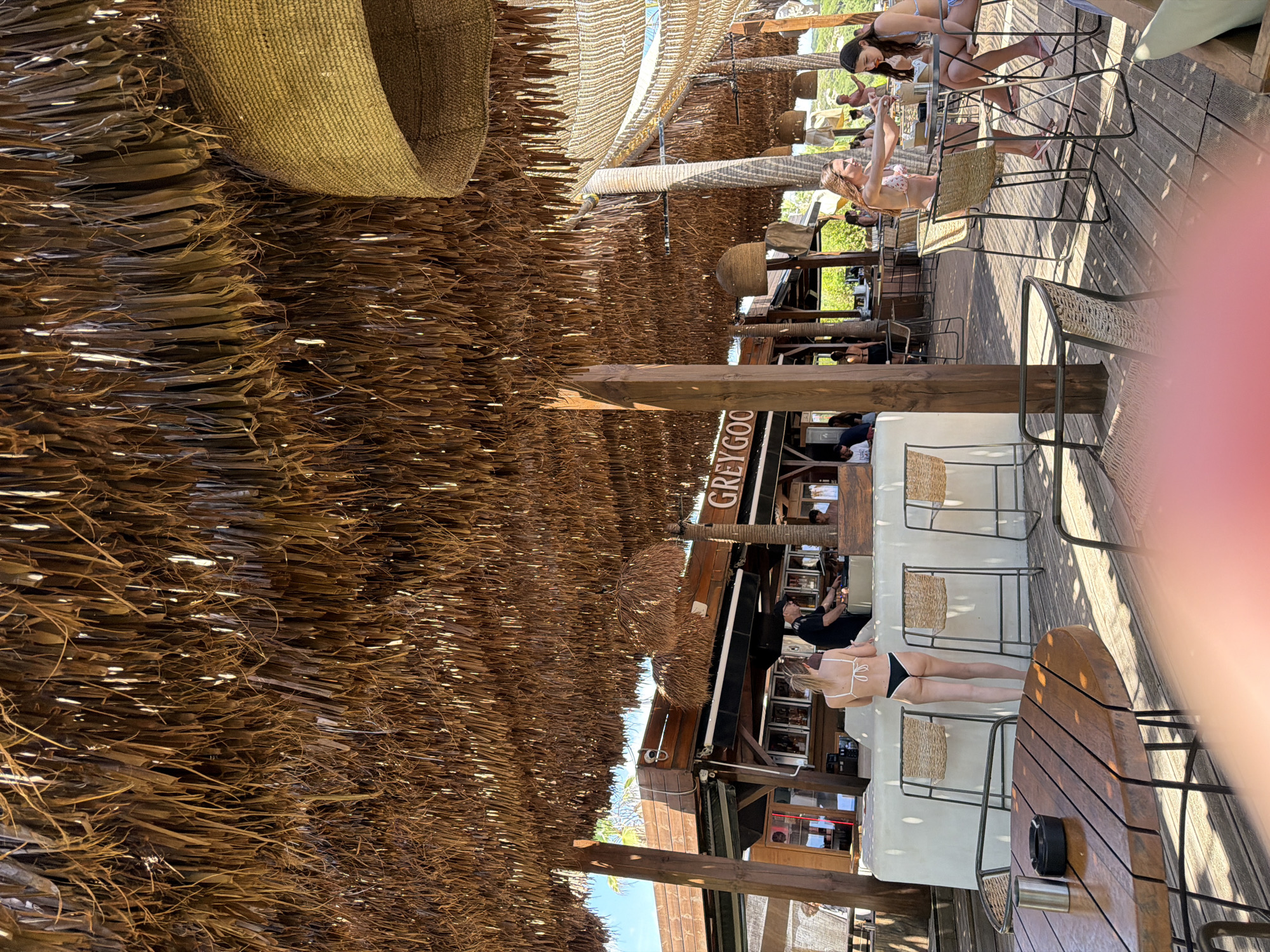 A beach bar in Nea Peramos, Greece, is shown from a low-angle perspective, looking up at a thatched roof. In the foreground, a round wooden table with metal legs is visible. Beyond the table, a white bar counter with stools is set up, where a person wearing a black hat and a dark shirt is preparing drinks. A woman in a white bikini stands facing the bar, her back to the camera. Further to the right, two women in bikinis are seated at high stools, engaged in conversation. The bar area is furnished with wooden floors and has a rustic, open-air design, with palm trees and vegetation visible in the background. The lighting suggests it is daytime, with sunlight filtering through the thatched roof.