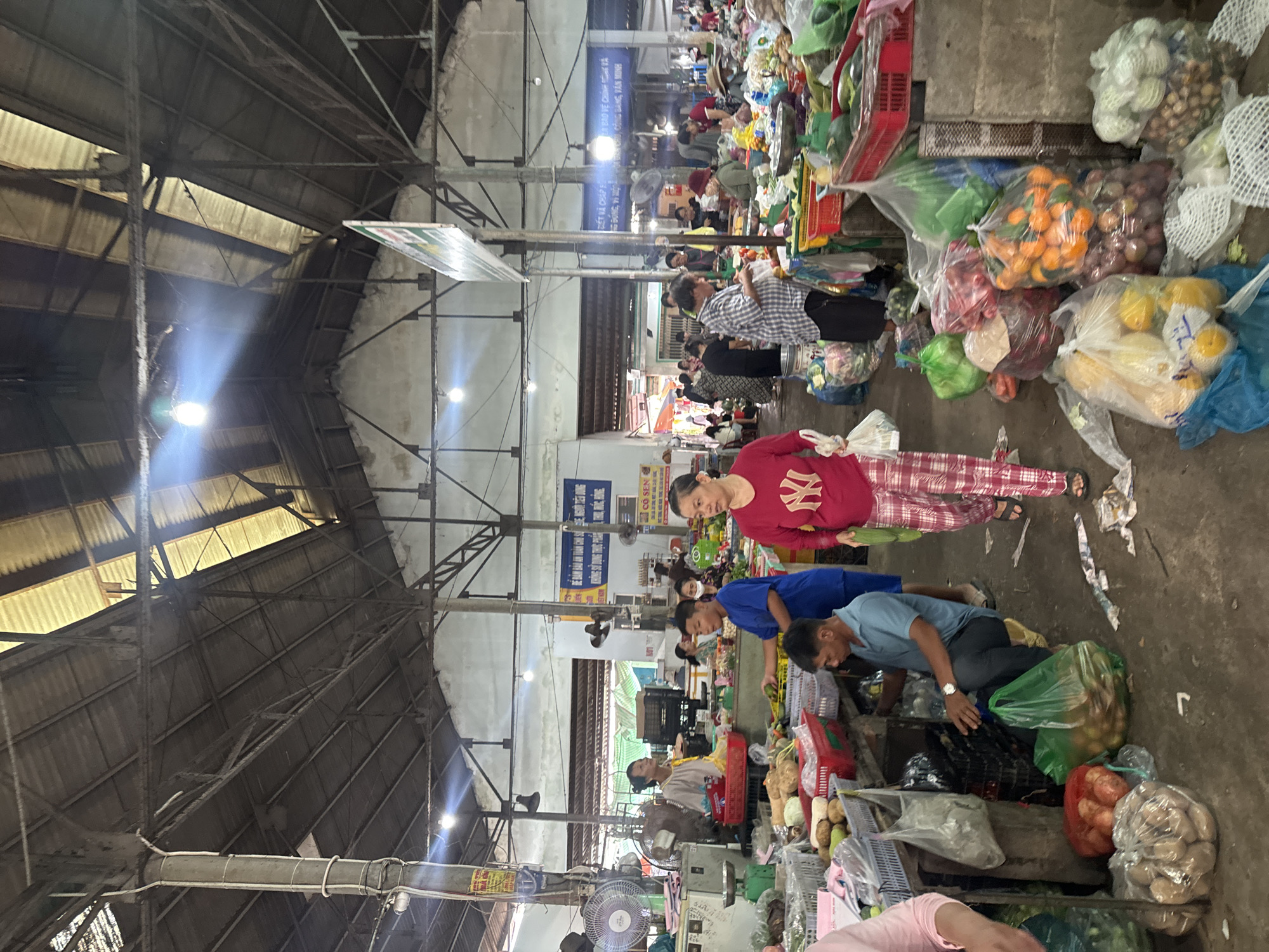 The image depicts the interior of a bustling marketplace in Mũi Né, Vietnam. The scene is characterized by a large, covered structure with a high, corrugated metal roof supported by metal beams and pillars. Natural light filters through gaps in the roof and from overhead bulbs. The ground is concrete, and the atmosphere is busy with people engaged in commerce.
Numerous stalls are visible, laden with a variety of fresh produce. Baskets and crates overflow with fruits and vegetables, including what appear to be potatoes, oranges, mangoes, and various leafy greens. Bags of produce are also piled on the ground. Customers and vendors are interacting, with some individuals examining goods and others attending to their stalls. The people in the image are dressed in casual clothing. Several individuals are wearing bright-colored shirts and pants, typical for a warm climate. Fans are visible in some areas, likely for ventilation. Signs in Vietnamese are posted on some of the stalls and structures. The overall impression is one of a lively and active local market.