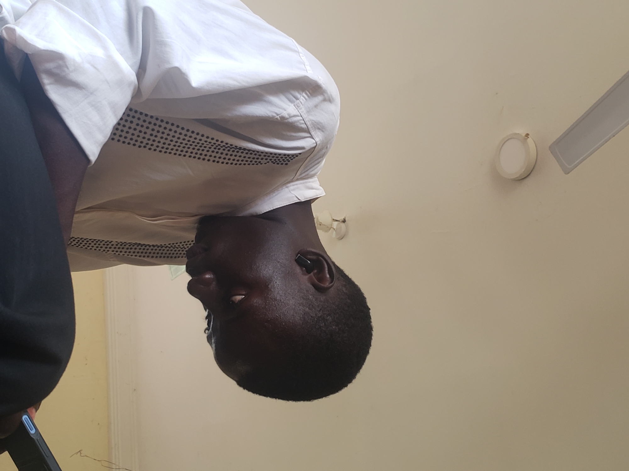 A young Black male, wearing a white long-sleeved shirt with black dotted trim, is seen from the chest up, looking down at a dark-colored smartphone in his hands. He has short black hair and is wearing a black earbud in his right ear. The background consists of a plain, off-white ceiling with a light fixture visible, and part of a ceiling fan. The image appears to be taken indoors, likely in a residential setting in Gumel, Nigeria, given the natural lighting and typical ceiling design.