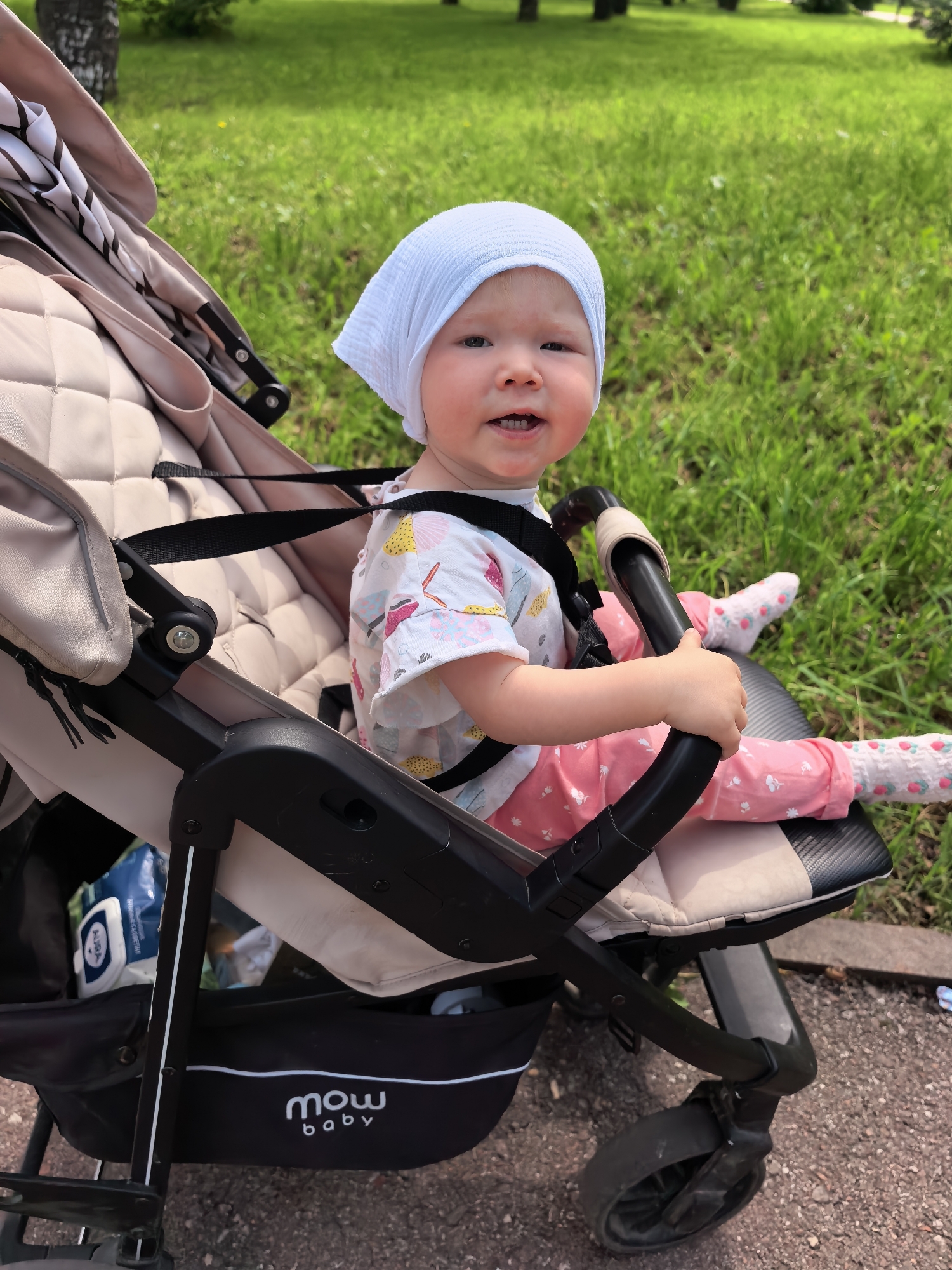 A young child wearing a light blue head covering, a white shirt with colorful abstract shapes, and pink pants with a floral pattern is sitting in a beige and black stroller. The child is looking at the camera with an open mouth and appears to be smiling. The child's feet are resting on the stroller's footrest, and they are wearing white socks with a strawberry pattern. The background is a grassy park with trees, suggesting an outdoor setting. The stroller has a black frame and beige padding, with a black harness secured around the child. The child is holding onto a black bar on the stroller. The scene is captured on a sunny day. This image was taken in the city of Sterlitamak, Russia.