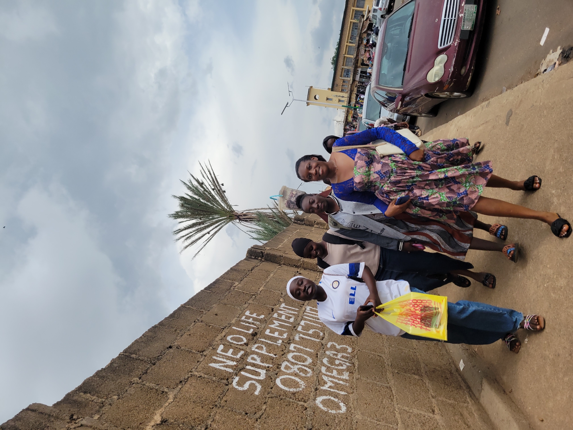 Four young women are walking along a sidewalk next to a concrete wall with white lettering. The woman in the front left is wearing a white t-shirt with blue and black trim, blue jeans, and flip-flops. She is holding a yellow plastic bag. The woman next to her is wearing a beige top and dark pants. The third woman from the left is wearing a gray jacket over a striped dress and dark pants, and carrying a beige purse. The woman in the front right is wearing a blue long-sleeved top and a patterned dress, and carrying a white purse. Behind them, there are cars and buildings visible. The sky is overcast. The lettering on the wall reads "NEO LIFE SUPPLEMENT 0807 7571111 OMEGA3."