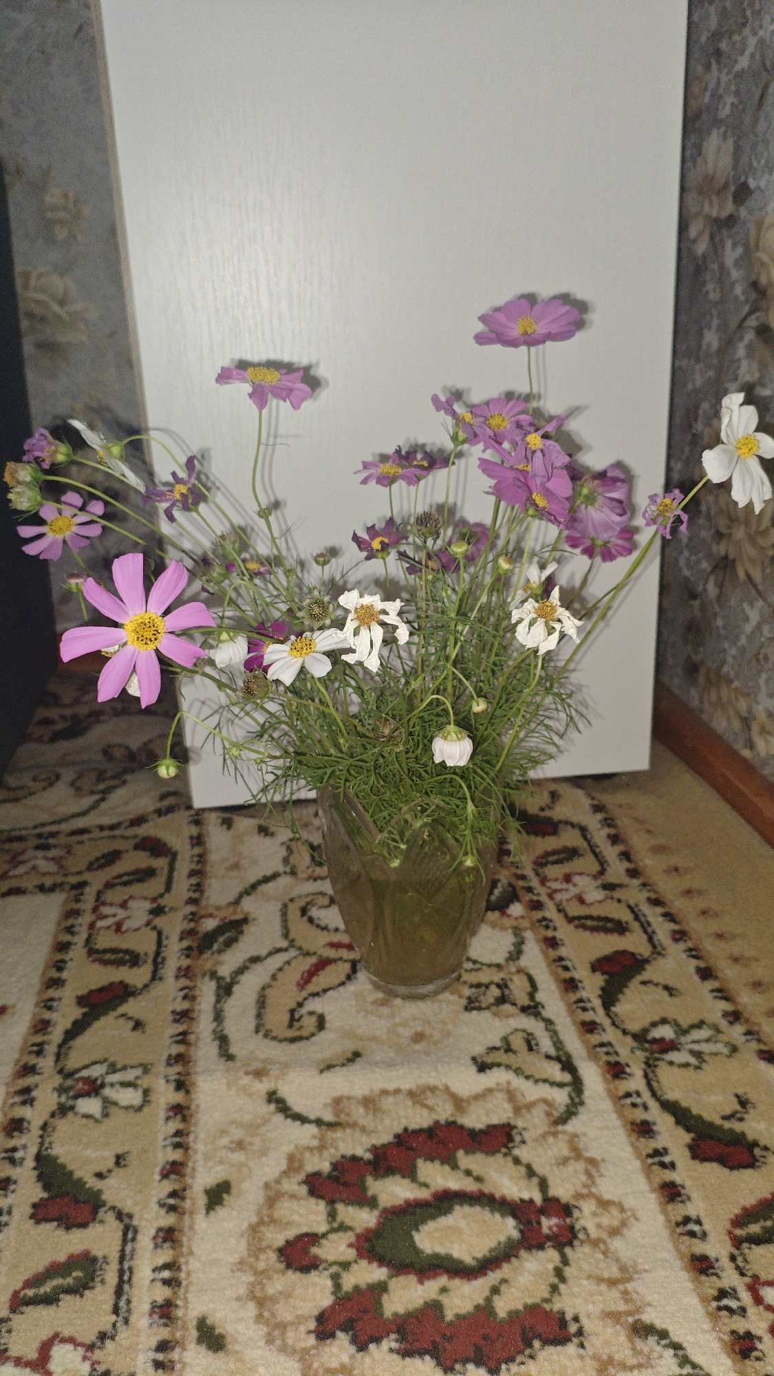 A bouquet of pink and white flowers is arranged in a clear glass vase. The vase sits on a beige carpet with a floral pattern. Behind the vase is a white door, and to the right of the door is a wall with floral wallpaper. The lighting in the room appears to be artificial.