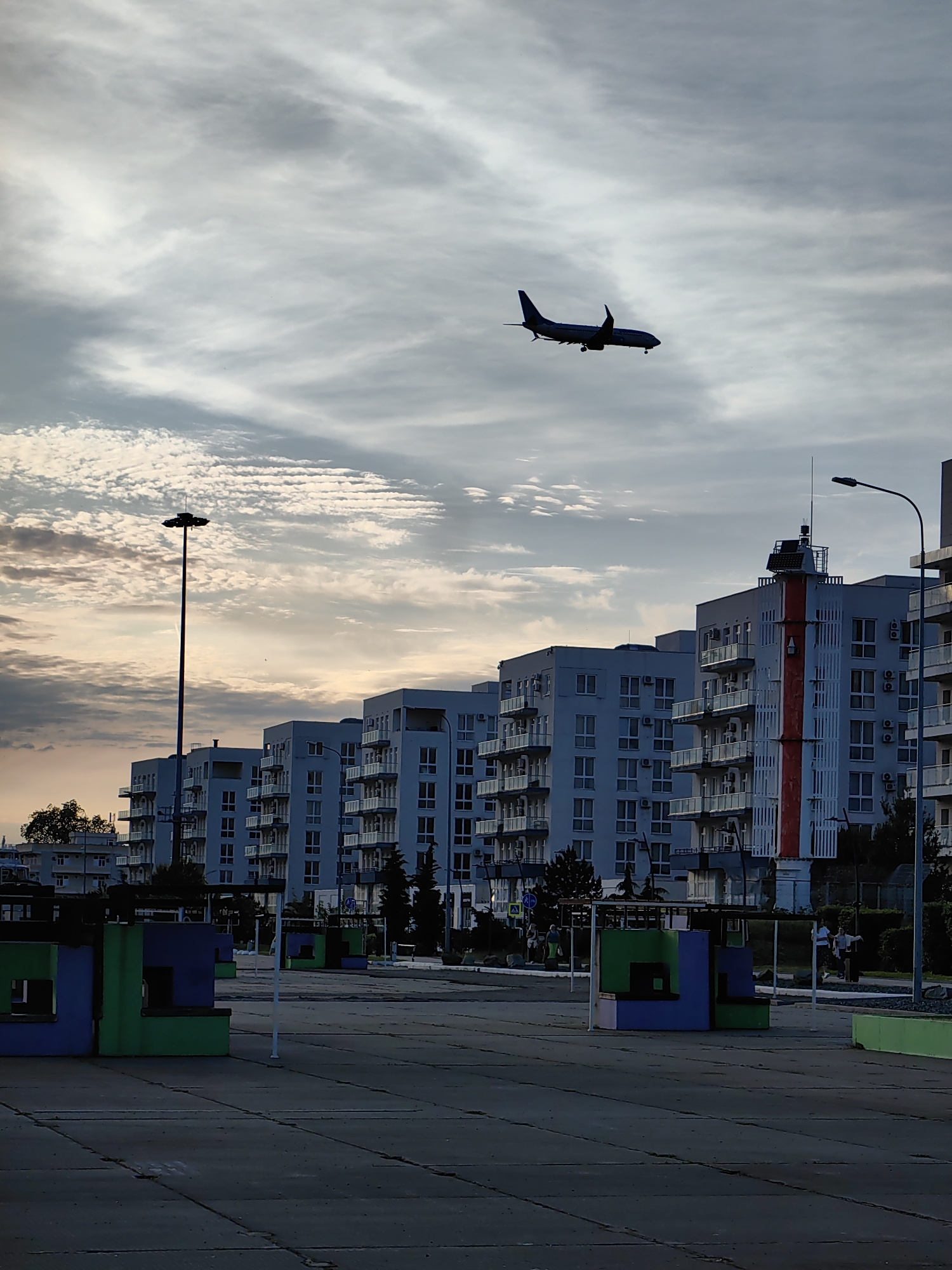 The image depicts an urban scene in Sirius, Russia, during what appears to be dusk or late afternoon, characterized by an overcast sky with patches of warmer light on the horizon. A large passenger airplane, silhouetted against the sky, is prominently featured in the upper middle, flying relatively low, suggesting it is either on approach to or departure from an airport.
Below the airplane, a row of modern, multi-story apartment buildings dominates the midground. These buildings are uniformly light-colored (possibly white or light blue) with numerous balconies and external air conditioning units. One building, towards the right, stands out with a striking vertical red and white stripe running down its facade, adorned with what looks like an antenna or sensor array on its roof. A tall, slender light pole with a large circular light fixture extends upwards from the left side, almost reaching the plane. Another street light is visible on the far right.
The foreground consists of a paved, dark gray surface, possibly a plaza or a large walkway, with visible seams. Scattered across this area are several colorful, geometric structures, rendered in shades of blue, green, and purple, possibly serving as benches or decorative shelters. In the background, behind the buildings, a few people are visible, engaged in casual activity, including one person on a bicycle on the right side. The overall atmosphere is one of contemporary urban life under a dramatic sky. No distinct text is visible.