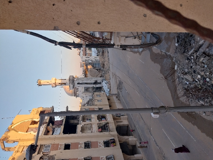 The image depicts a scene of extensive urban destruction in Khan Yunis, Palestinian Territory, captured from an elevated perspective under a clear, pale blue sky at what appears to be either dawn or dusk. In the immediate foreground on the right, a textured beige building wall displays multiple small impact marks, with several bundles of dark cables attached to it. A twisted piece of metal rebar is visible in the extreme lower right. Below, a substantial pile of concrete rubble, rebar, and assorted debris indicates severe structural collapse.
A grey asphalt road with wet patches runs through the midground. A tall, grey lamppost is centrally positioned on the left side of the road, partially obscuring a damaged building. An individual dressed in a dark red abaya and head covering walks leftwards on the road. Further left, three small figures, likely children, stand on the roadside adjacent to a damaged multi-story building.
The central background is dominated by a severely damaged mosque, identifiable by its large dome and a largely intact, tall cylindrical minaret. Extensive structural breaches are visible on the mosque, with significant portions of its walls and roof destroyed, exposing its interior and surrounding it with debris. To the left of the mosque, the multi-story building exhibits severe damage, including partial collapse of upper floors, exposed rebar, and blackened sections suggestive of fire. Balconies with clothing hanging are present on its remaining levels. Further damaged structures extend into the distance, indicating widespread devastation across the urban landscape.