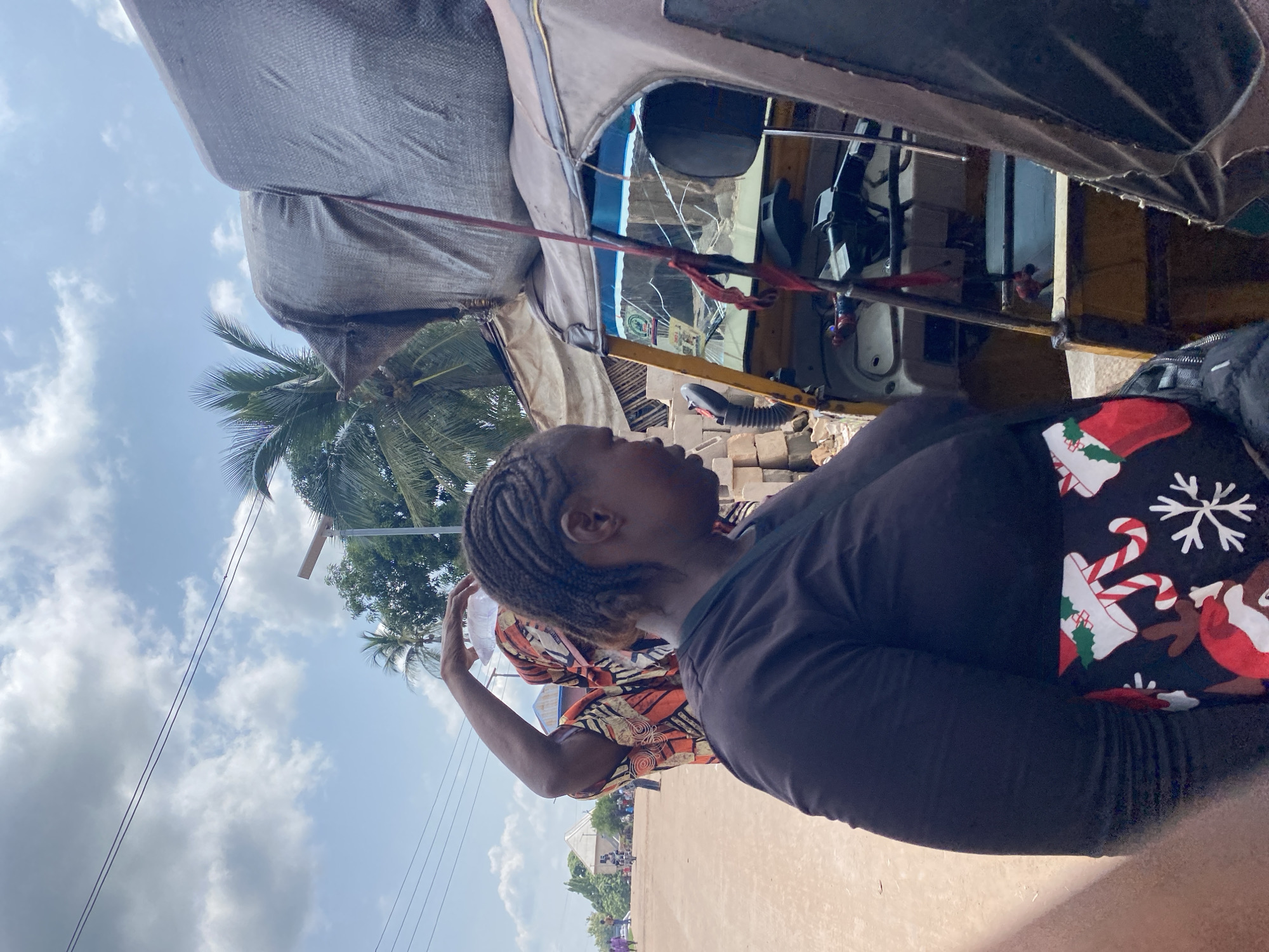 An outdoor scene in Nguzu Edda, Nigeria, depicts a woman from the chest up, facing right and looking down. She has dark skin and braided hair, wearing a dark long-sleeved shirt and a black skirt with a Christmas-themed pattern of snowflakes, reindeer, and candy canes. To her left, another person, also with dark skin and braided hair, is visible from the shoulders up. This person is wearing an orange, yellow, and black patterned garment and has their right arm raised, holding a clear plastic container on their head. They are looking to their left. In the background, a yellow and tan-colored auto-rickshaw, also known as a "keke," is parked. The vehicle is partially covered with a large, grey, textured tarp. A red ribbon or cord hangs from a pole near the auto-rickshaw's windshield. The ground is a dusty, light brown surface, and the sky above is blue with scattered white clouds. Palm trees and other vegetation are visible in the distance, along with some buildings and other people walking or on bicycles.