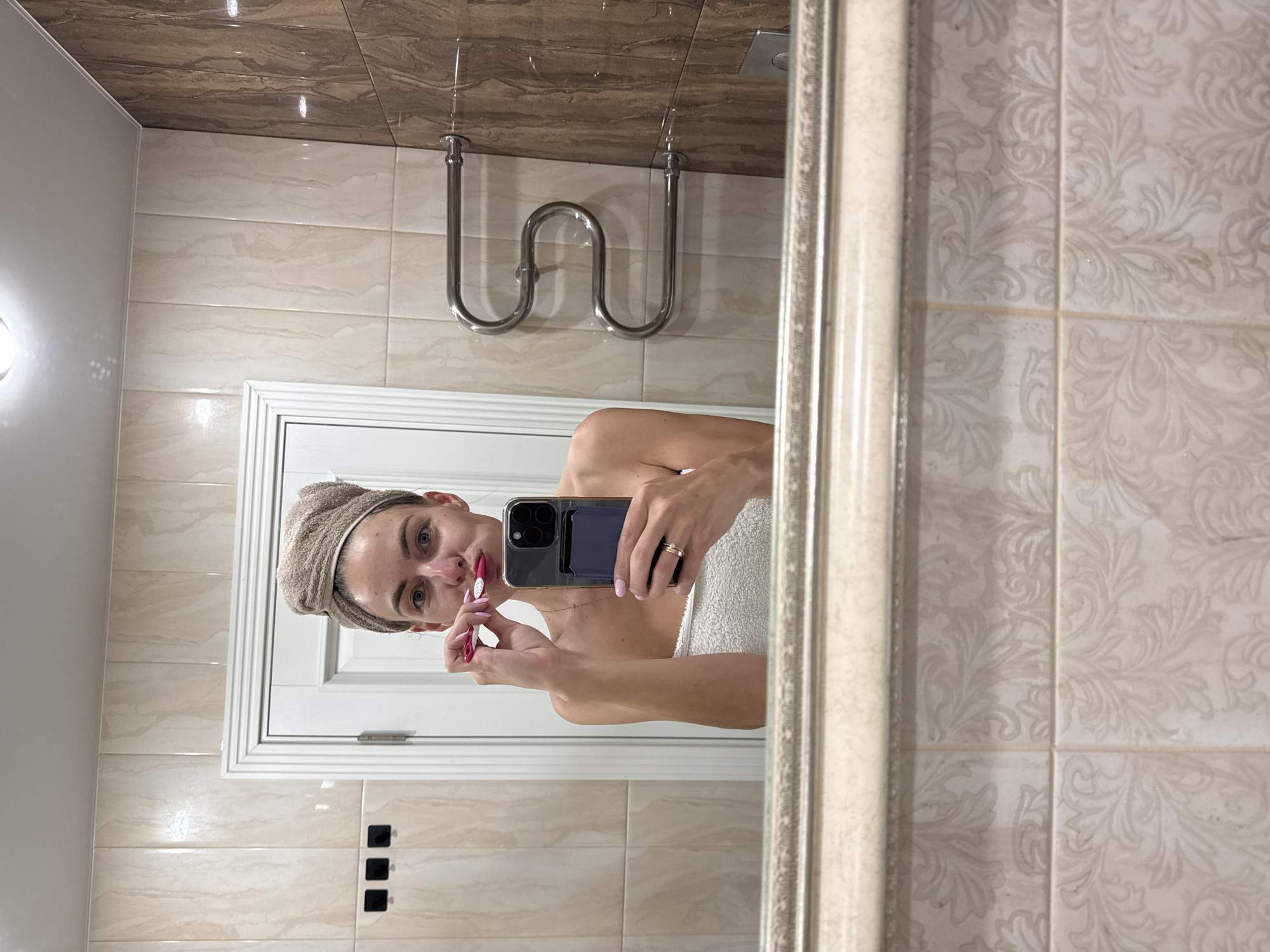 A woman is captured in a mirror selfie in a bathroom in Sochi, Russia, as she brushes her teeth. She is wearing a towel wrapped around her body and another towel on her head. Her face is visible, and she is holding a pink toothbrush and her smartphone. The bathroom features tiled walls with a marble-like pattern and a chrome towel rack. The mirror is framed with ornate detailing.