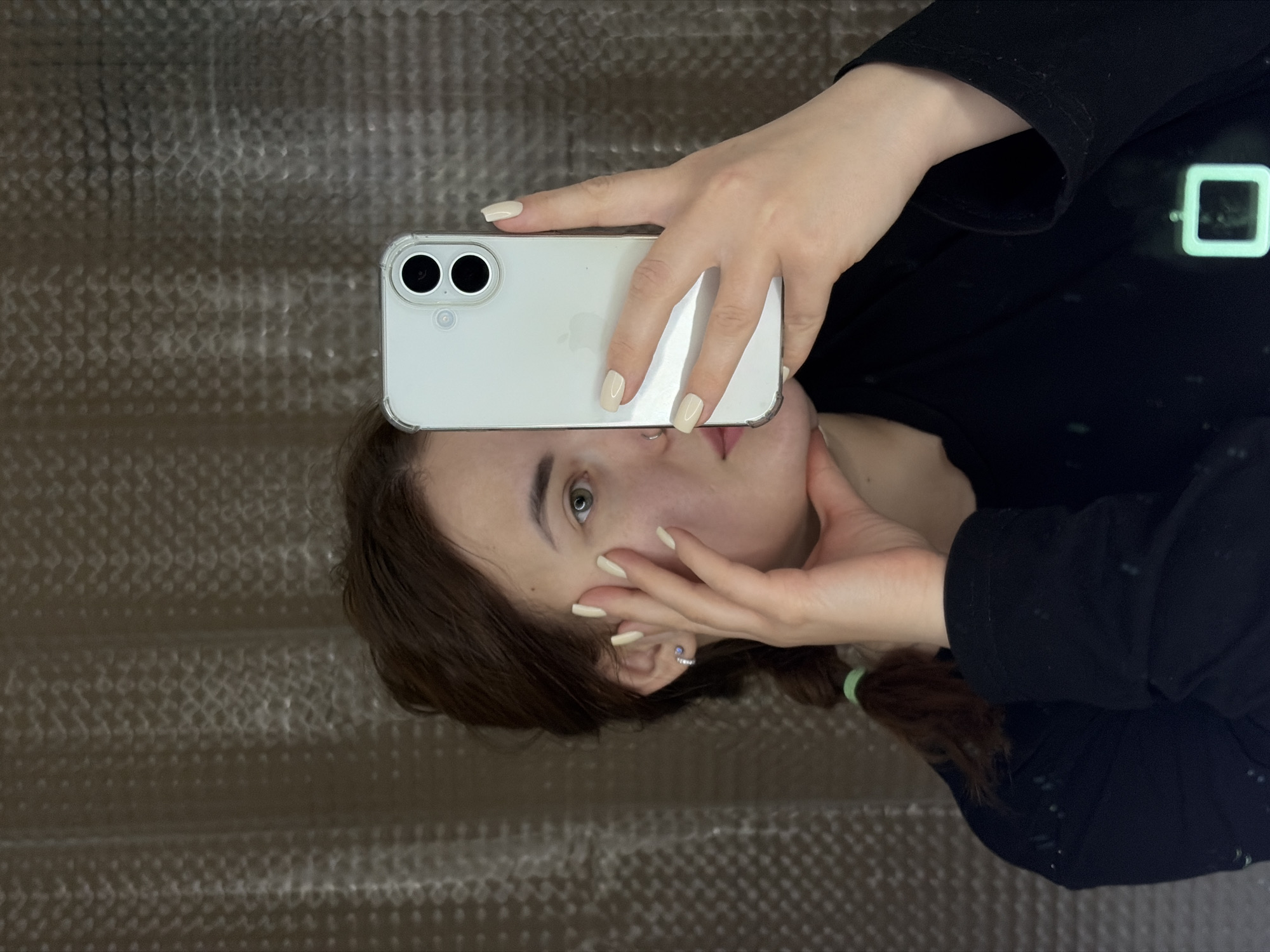 A young woman with light skin is taking a selfie with a white iPhone 11. The phone has a clear case. She is wearing a black long-sleeved shirt. Her hair is brown and pulled back in a ponytail with a green hair tie. She has pale yellow nail polish. She is holding the phone with her left hand and touching her face with her right hand. The background appears to be a textured wall. There is no visible evidence to indicate that the photo was taken in Krasnodar, Russia.