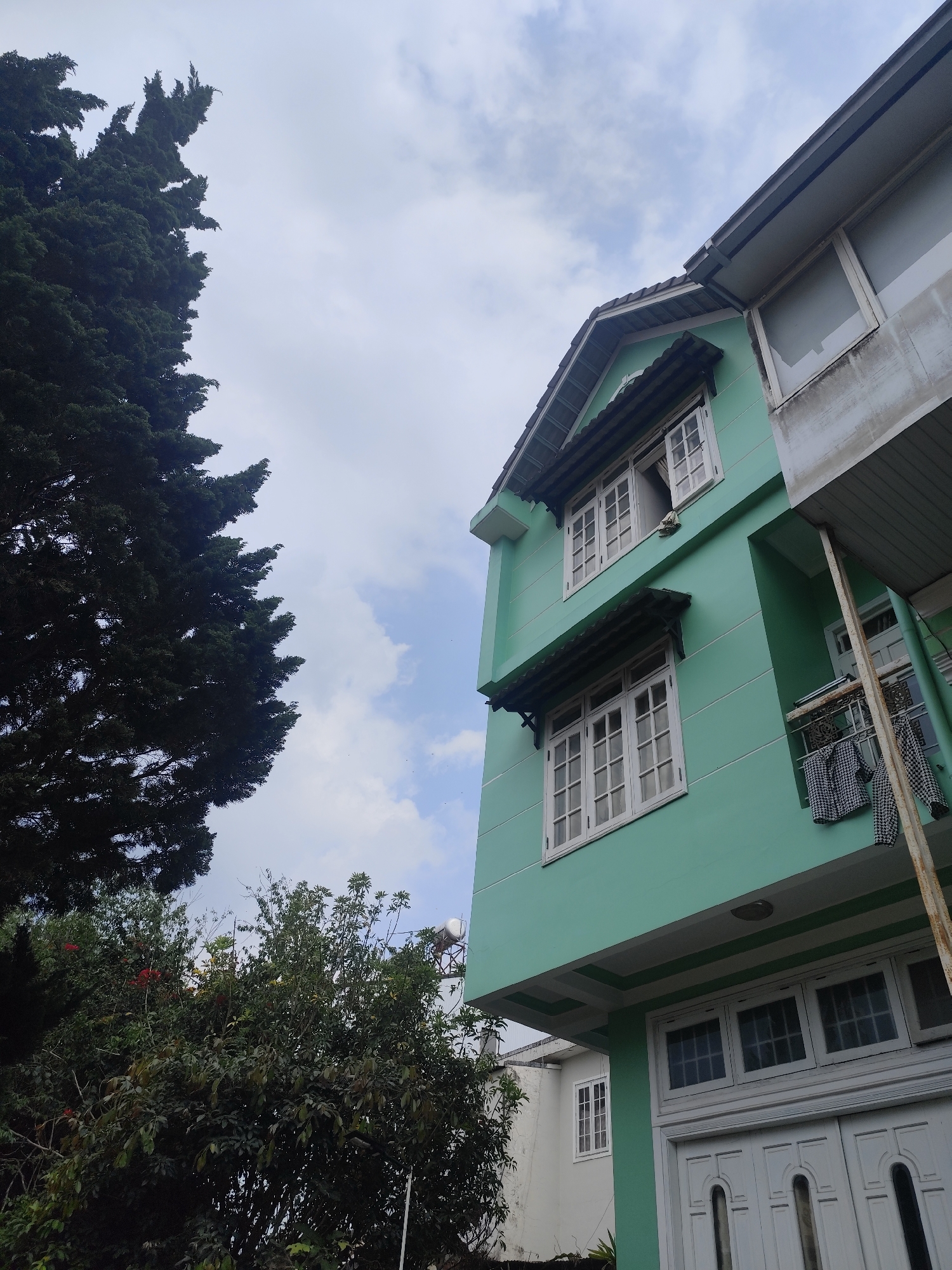 This image presents a low-angle view of a multi-story house in Da Lat, Vietnam, framed by lush greenery and a cloudy sky. The house is painted a distinct light mint green, featuring multiple white-framed windows. On the second floor, one window is open, and another lower window is covered by a dark tiled awning. The house has a dark tiled roof with visible gutters. To the right, an adjacent section of the building, possibly an extension, is unpainted and appears greyish-white, with a large window and a small balcony-like area where black and white patterned clothes are hanging to dry.
A prominent, tall evergreen tree with dense, dark green foliage occupies the entire left side of the frame, reaching towards the sky. Below the house, further green bushes and trees are visible, with hints of red flowers adding a touch of color. In the background, partly obscured by foliage, a white cylindrical object, likely a water tank on a stand, can be seen.
The sky is overcast with a mix of white and light blue clouds, suggesting daytime without direct sunlight. There are no people visible, and the scene depicts a quiet, residential setting. No text is discernible within the image. The overall impression is a serene and green environment characteristic of Da Lat.