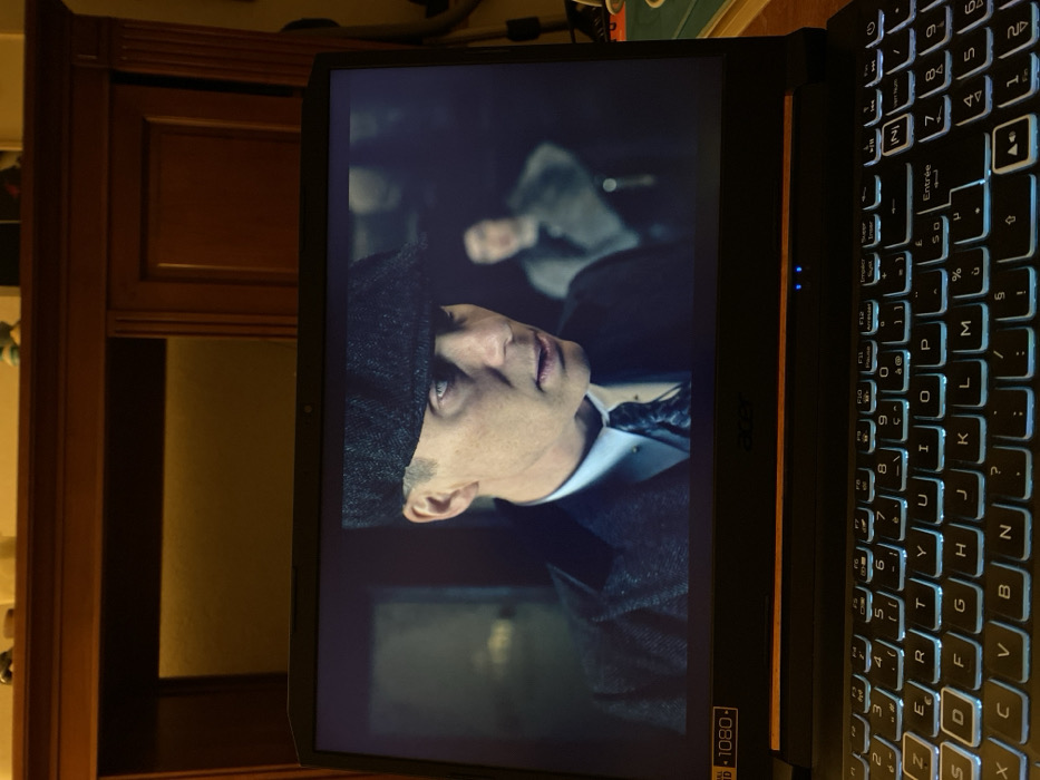 This image captures a black Acer laptop displaying a scene from a television series or film, positioned indoors in Bonnieux, France. The laptop features a backlit QWERTY (French AZERTY) keyboard with light blue illumination and a screen bezel indicating "1080p" resolution. The screen shows a male character, identified as Cillian Murphy's character Thomas Shelby from "Peaky Blinders," prominently in the foreground. He wears a dark tweed flat cap, a dark overcoat, a white collared shirt, and a patterned tie with a tie pin. His expression is intense, with a direct, serious gaze. A second, blurry individual, possibly female and wearing a light-colored, fur-collared garment, is visible in the darker background to the right of the primary character. Behind the laptop, a substantial piece of dark brown wooden furniture, possibly a cabinet or hutch, occupies the upper portion of the frame. It features carved panels and is dimly lit, with a warm, yellowish glow emanating from behind recessed sections. Small, indistinct objects are present on the top surface of the furniture. The overall environment suggests an indoor setting with low ambient lighting, focusing on the screen's content.