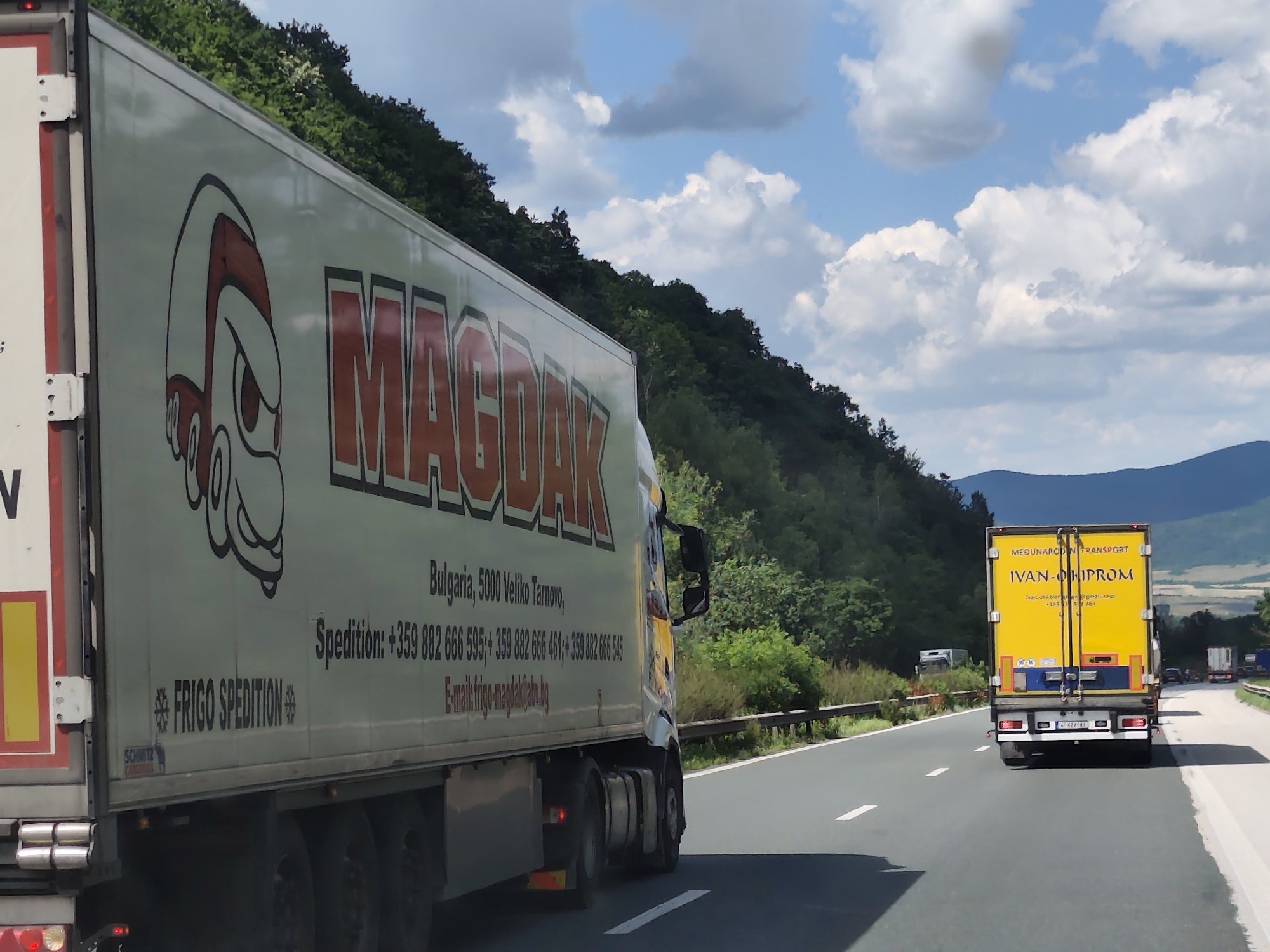 This media file depicts a daytime scene on a highway, likely in Bulgaria, given the mention of "Veliko Tarnovo." The view is from a vehicle moving along the road, focusing on the side of a large, white semi-trailer truck on the left. This truck features prominent red lettering that reads "MAGDAK" and a cartoonish illustration of a character's head with red and white stripes. Below the company name, text indicates "Bulgaria, 5000 Veliko Tarnovo," and contact information including a phone number "+359 882 666 595" and an email address "E-mail: frigo-magdak@abv.bg". The words "FRIGO SPEDITION" are also visible on the truck's side.
In the distance, another yellow semi-trailer truck is visible, also traveling on the highway. This truck has the text "MEDUNARODNI TRANSPORT" and "IVAN-OHIPROM" in blue lettering on its back. The highway itself is paved and has white lane markings. The surrounding landscape is characterized by a steep, tree-covered hillside to the left and a more distant, rolling mountainous terrain under a sky filled with white, fluffy clouds, suggesting fair weather. The overall impression is of a journey on a sunny day.