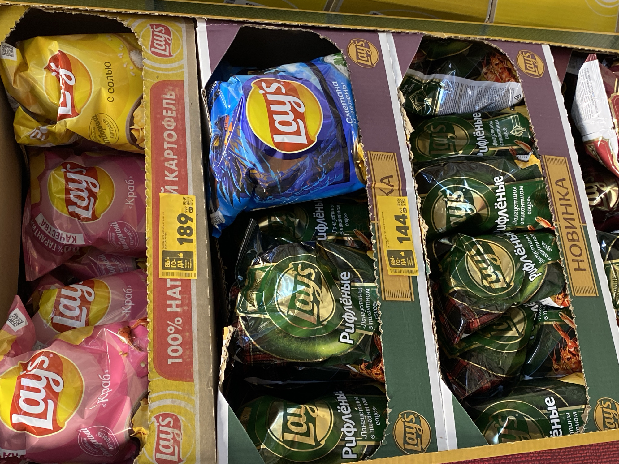 The image depicts a supermarket shelf in Votkinsk, Russia, stocked with Lay's potato chips. Three different flavors are visible, arranged in cardboard display cases: "Crab" and "Salt" on the left, "Sour Cream and Herbs" in the center, and "Lobster in Spicy Sauce" on the right. Price tags indicate the cost of each flavor: 189.99 for "Crab", and 144 for "Lobster in Spicy Sauce". The product is arranged in a way that is easy to reach and read. The colors of the packaging are vibrant and appealing, with pink for "Crab", blue for "Sour Cream and Herbs", and green for "Lobster in Spicy Sauce". The cardboard display cases feature the Lay's logo and branding. The "Lobster in Spicy Sauce" packaging has a "New" banner on it.