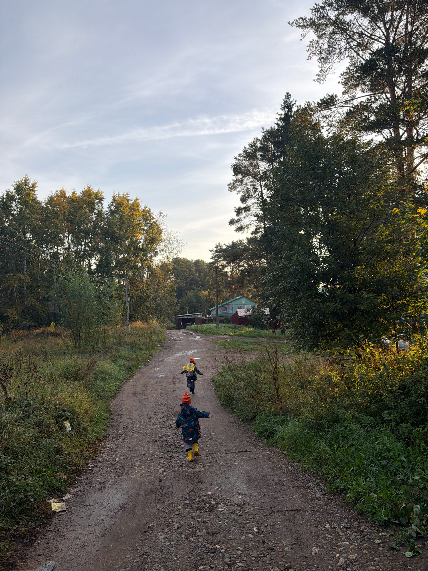 Two children, one in front wearing a dark blue jacket with a dinosaur pattern and yellow boots, and another behind wearing a yellow backpack and orange hat, are running up a dirt road. They are in a rural or semi-rural area with trees on either side of the road. A light blue house with a red foundation is visible in the distance. The sky is cloudy and the sun appears to be low in the sky, casting a warm light on the trees. The road is uneven and appears to have puddles. The overall impression is one of a casual, outdoor activity in a natural setting.