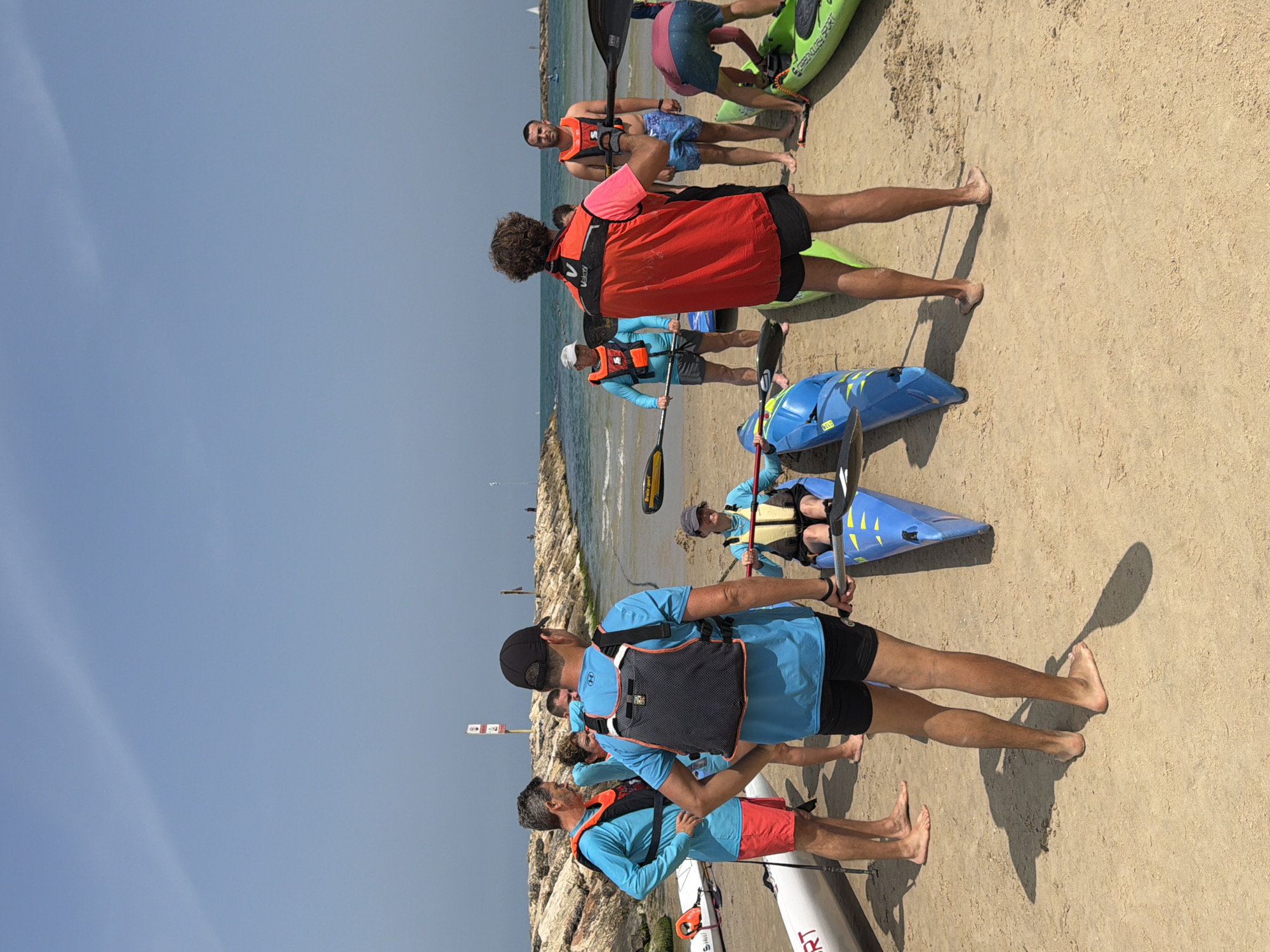 The image captures a group of people on a sandy beach in Tel-Aviv, Israel, preparing for or having just completed a kayaking activity.
People: There are several individuals, both men and women, wearing life vests. Some are holding paddles, suggesting they are involved in water sports. Some are barefoot.
Objects: Kayaks of varying colors (green, blue, and white) are scattered on the beach. Paddles are present, either held by individuals or near the kayaks. A black backpack is worn by one of the people.
Actions: The people are standing, some appearing to be in conversation or receiving instructions. Others are near the kayaks, presumably preparing to launch or disembark.
Interactions: Individuals are interacting with each other and with the equipment (kayaks and paddles).
Environment: The scene is set on a sandy beach with the Mediterranean Sea visible in the background. There are some rocks or a breakwater visible in the water. The sky is clear and blue.