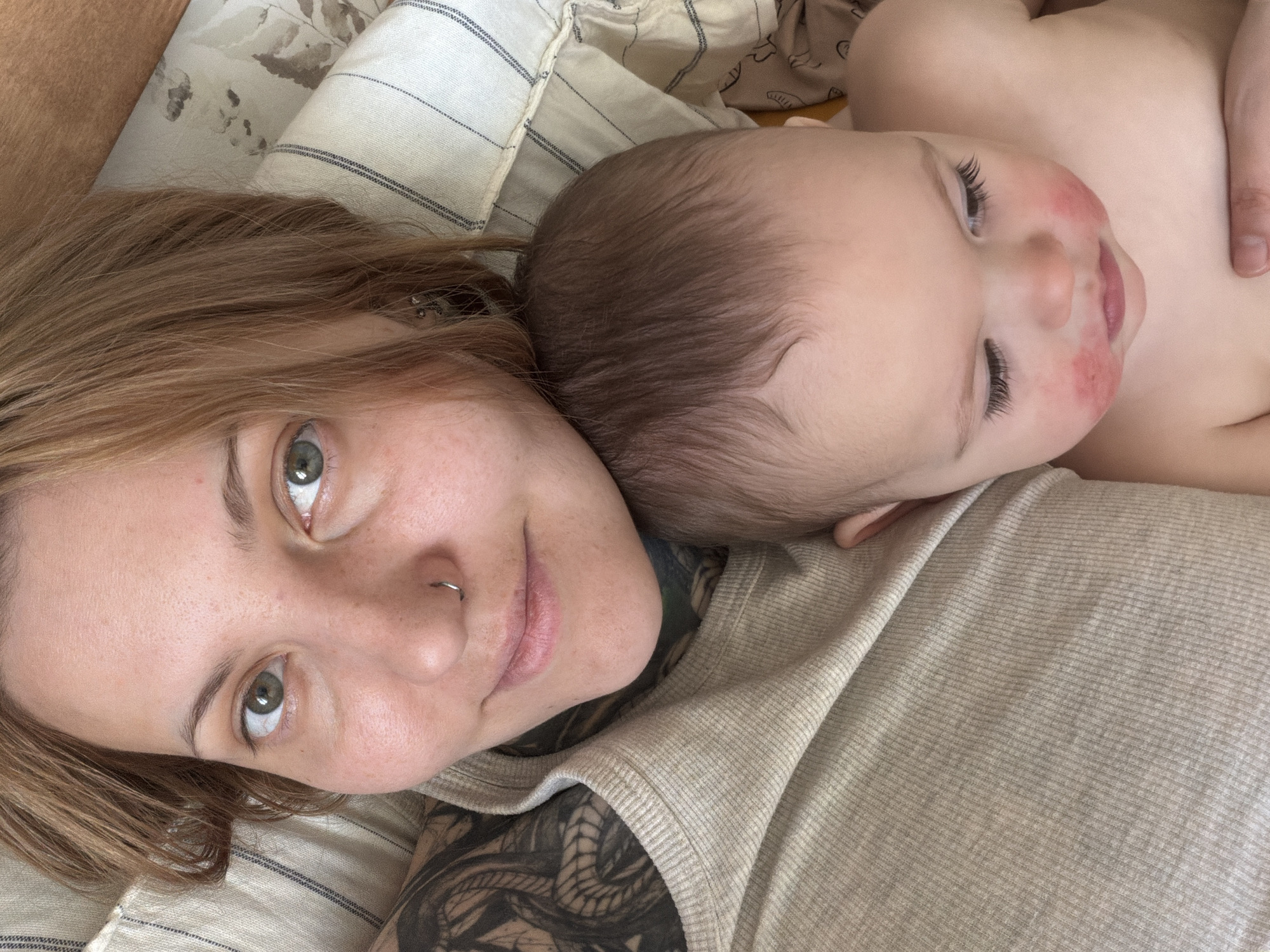 A close-up shot shows a woman lying on her back with a baby resting on her chest. The woman has light brown hair, green eyes, a nose ring, and tattoos on her arm. The baby appears to be sleeping, with some redness on its cheeks. They are both lying on a striped fabric surface, possibly a bed or couch. The location is unknown, and there is no indication of Chaikovsky, Russia.