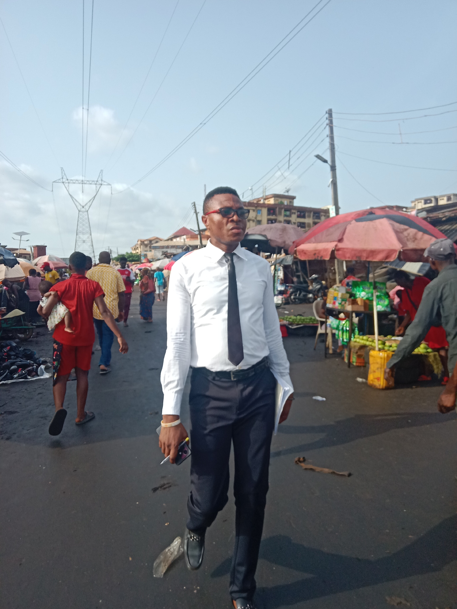 The image shows a man dressed in a white long-sleeved shirt, black tie, and black pants walking in a busy outdoor market. He is wearing dark-rimmed glasses and carrying a phone and possibly a pen or cigarette in his left hand. People are walking around him, and market stalls with red umbrellas are visible in the background, displaying various goods including green fruits or vegetables. The road or ground appears wet and potentially littered with debris. An electrical power line tower is visible in the background.