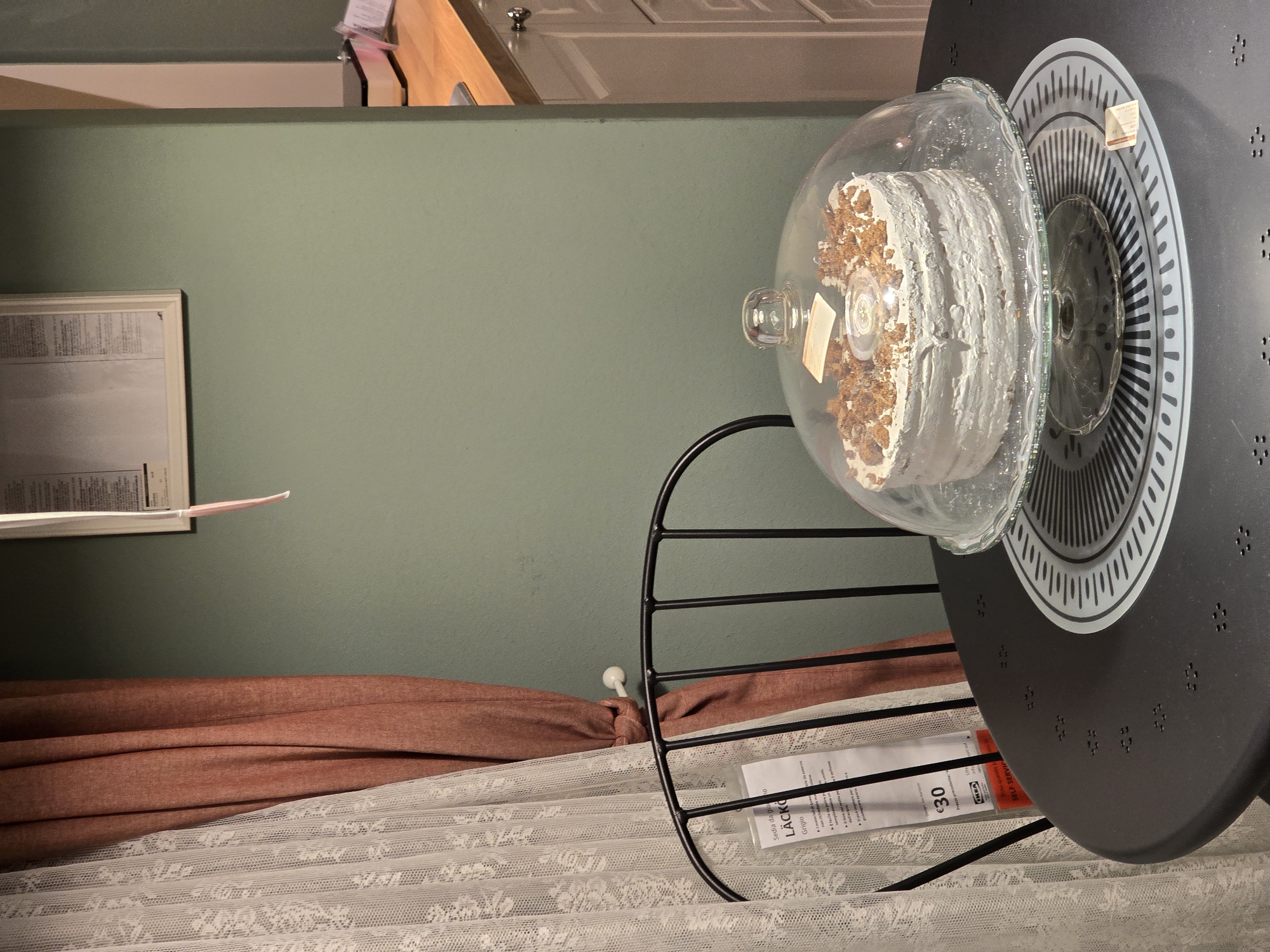 A round, white cake topped with crumbled pastry sits on a glass cake stand under a clear glass dome. The cake stand rests on a circular placemat with a black and white patterned design, which is placed on a dark round table. In the background, a peach-colored curtain is drawn to the left of a white lace curtain. A metal chair is partially visible to the left of the table. A poster is mounted on a light green wall behind the chair and curtains. To the right, a wooden countertop with white cabinets is visible. The scene appears to be in a domestic setting, potentially a dining room or kitchen, within a residential building in Baronissi, Italy.