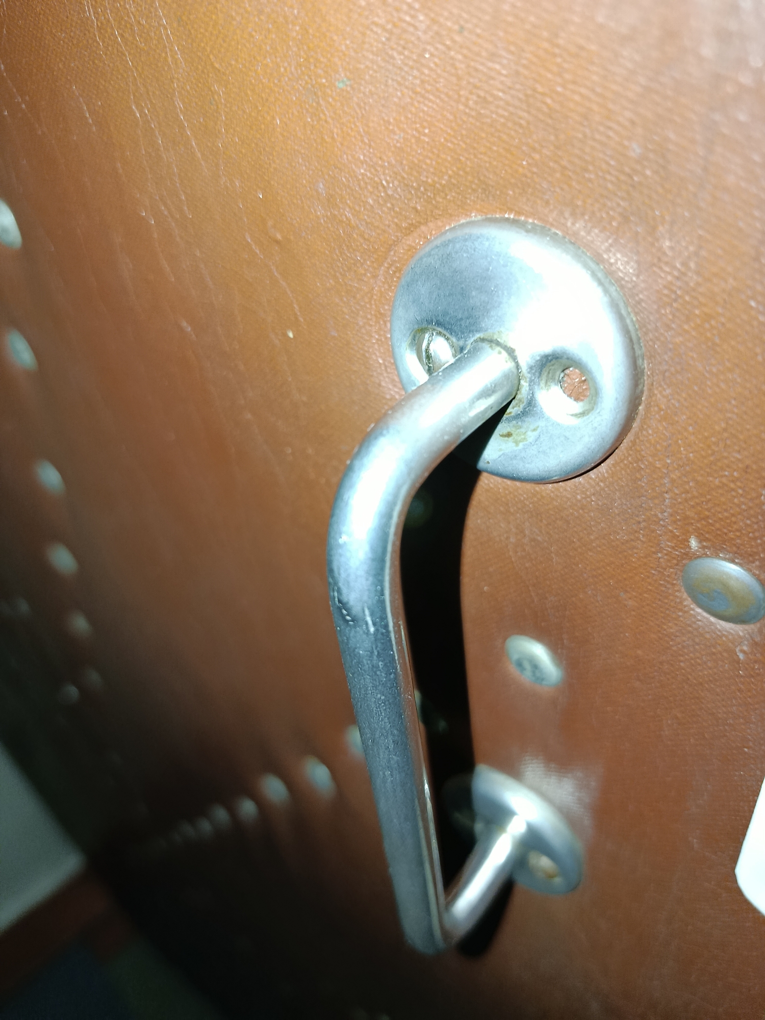 The image displays a close-up, oblique view of a metal handle attached to a brown, textured surface, possibly leather or a similar material. The handle is curved and polished, showing signs of wear and minor discoloration. It is secured by two round, flat metal escutcheons, each with two visible screw holes. The surface has a repetitive pattern of small, raised dots or rivets arranged in rows, some of which are partially visible in the lower left and right portions of the frame. The lighting creates shadows and highlights, emphasizing the textures and contours of the objects. There are no people visible in the image. The location of this object within the city of Tashkent, Uzbekistan, cannot be determined from the visual information provided.