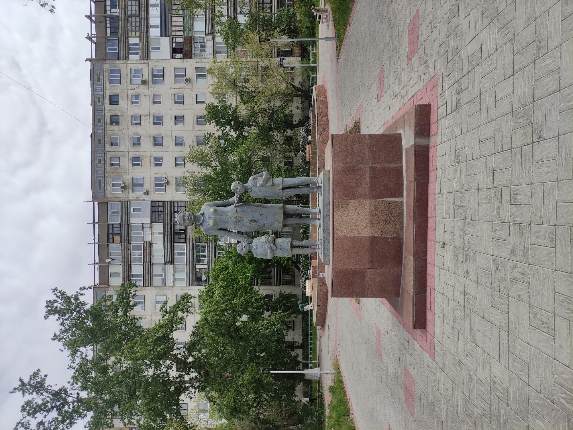 A bronze statue depicts a woman, a boy, and a girl standing on a brown stone pedestal. The woman stands in the center, the boy is on her right, and the girl is on her left. The statue is located on a paved walkway with red accents. Behind the statue, there is a tall apartment building with many windows, interspersed with green trees. In the background to the right, there is a white street lamp and a bench. This is the monument to the Mother in the city of Pavlodar, Kazakhstan. The sky is overcast.