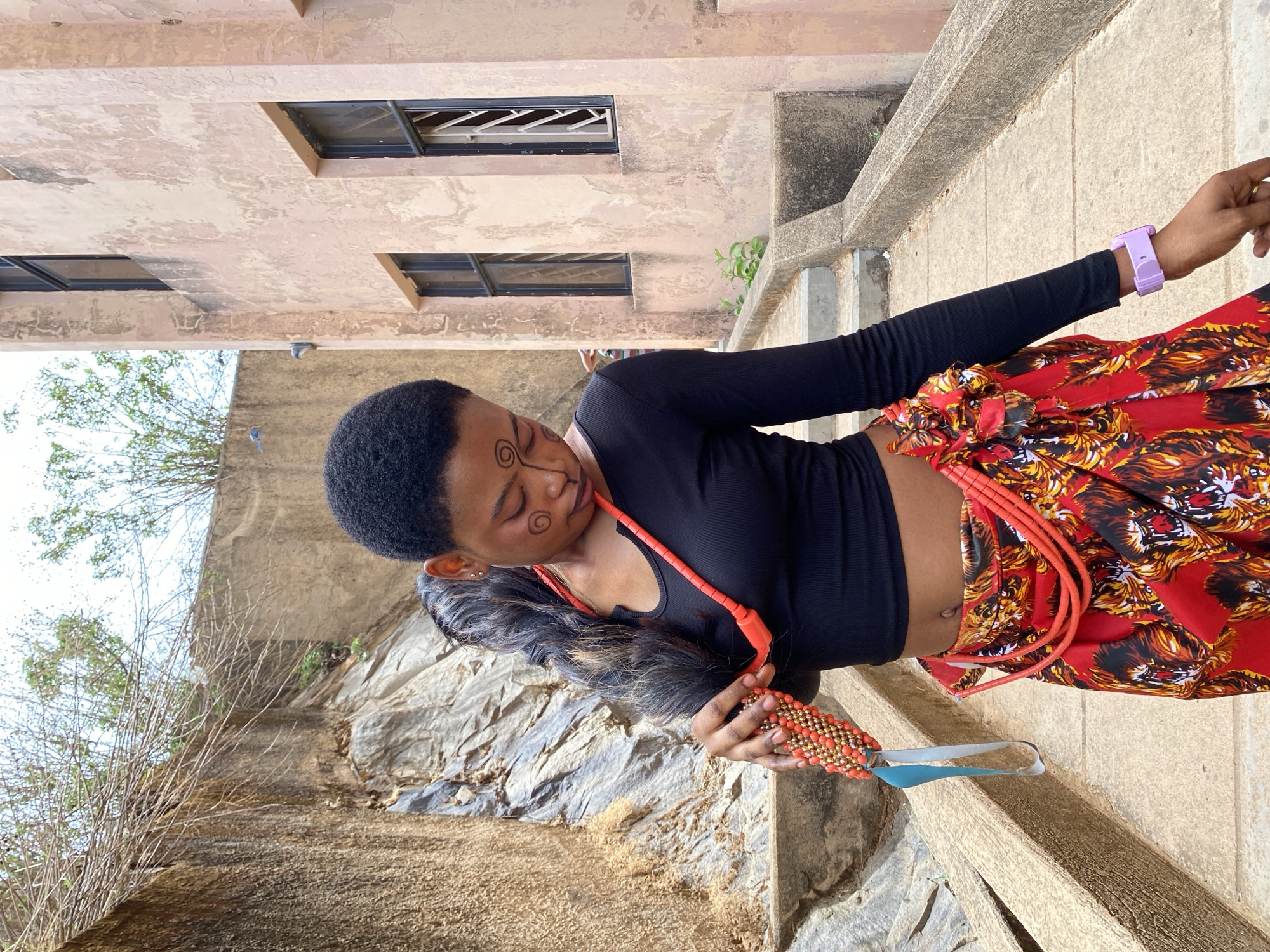 A woman stands outdoors in Gwong, Nigeria. She is wearing a long-sleeved black top, a vibrant red skirt, and a purple wristband. The woman has short black hair and tribal face paint. She is holding a beaded object with a blue ribbon. Behind her, a weathered building is visible, and a rocky landscape is to the left.