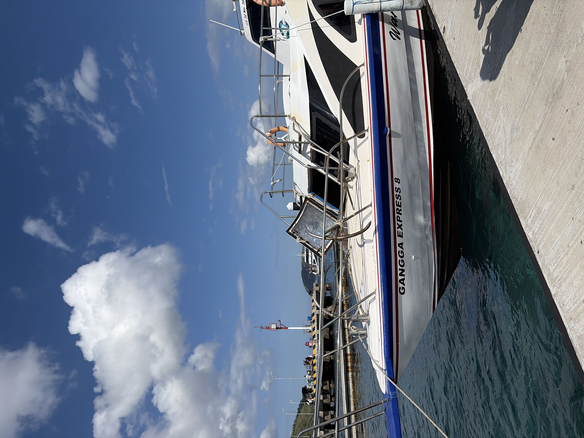 The image shows a white boat with black and blue accents docked at a pier in Kampung Toyapakeh, Indonesia. The boat is named "GANGGA EXPRESS 8" in black lettering. The pier is made of concrete and is lined with bollards. There are several people on the pier, some of whom appear to be waiting to board or disembark the boat. The water is a clear turquoise color. In the background, there are lush green hills and a bright blue sky with fluffy white clouds. The scene is peaceful and serene, with a sense of calm and tranquility. The overall impression is one of a beautiful day at a tropical paradise.