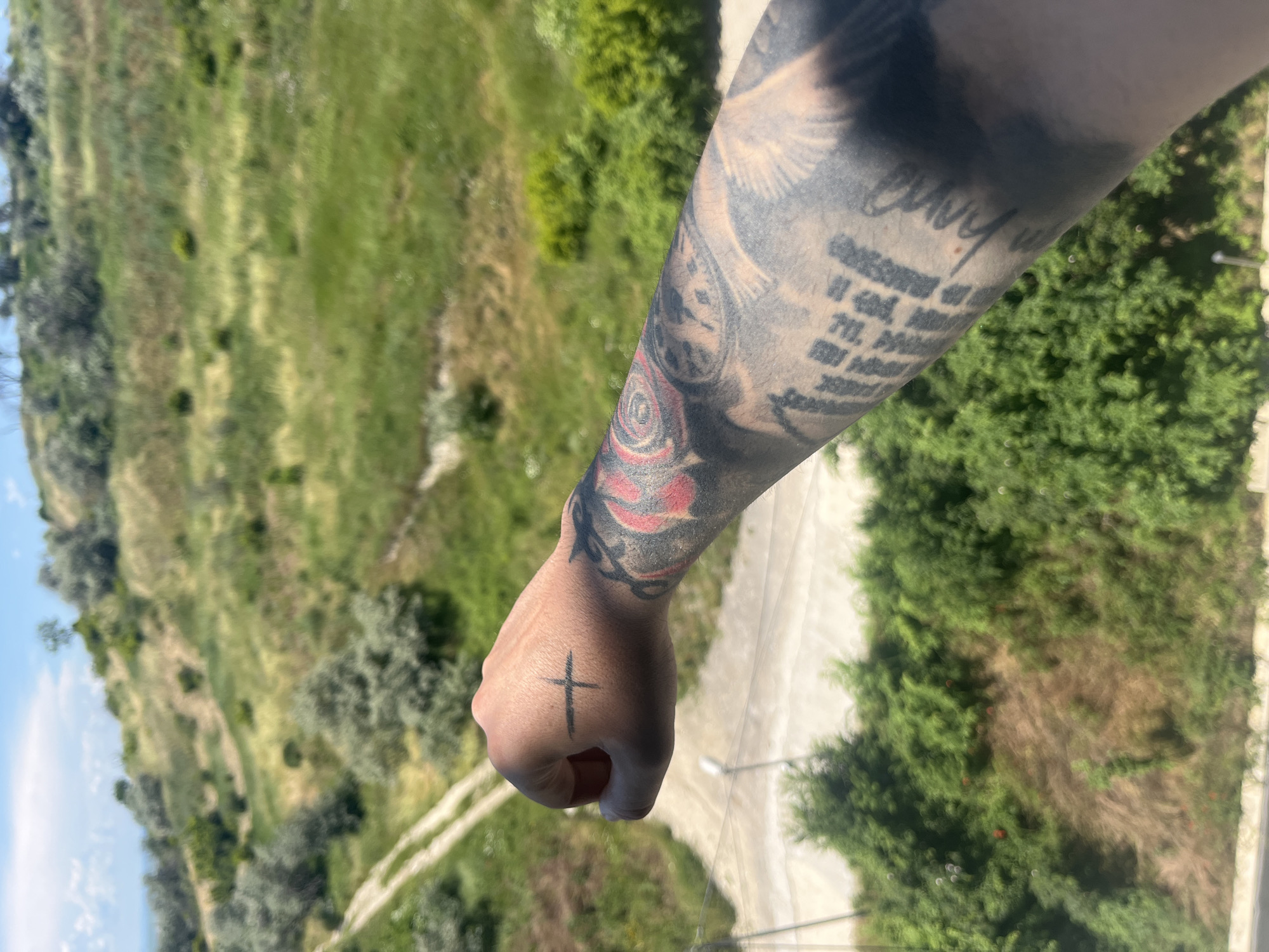 A tattooed arm is extended towards a grassy, hilly landscape with scattered trees and a dirt path. The arm displays a cross tattoo on the back of the hand and extensive black and red ink sleeves on the forearm, including a rose design. The background features a bright sky with some clouds and lush green vegetation, indicative of a warm, sunny day. The scene appears to be an outdoor location in Varna, Bulgaria, likely an elevated position overlooking a natural area.