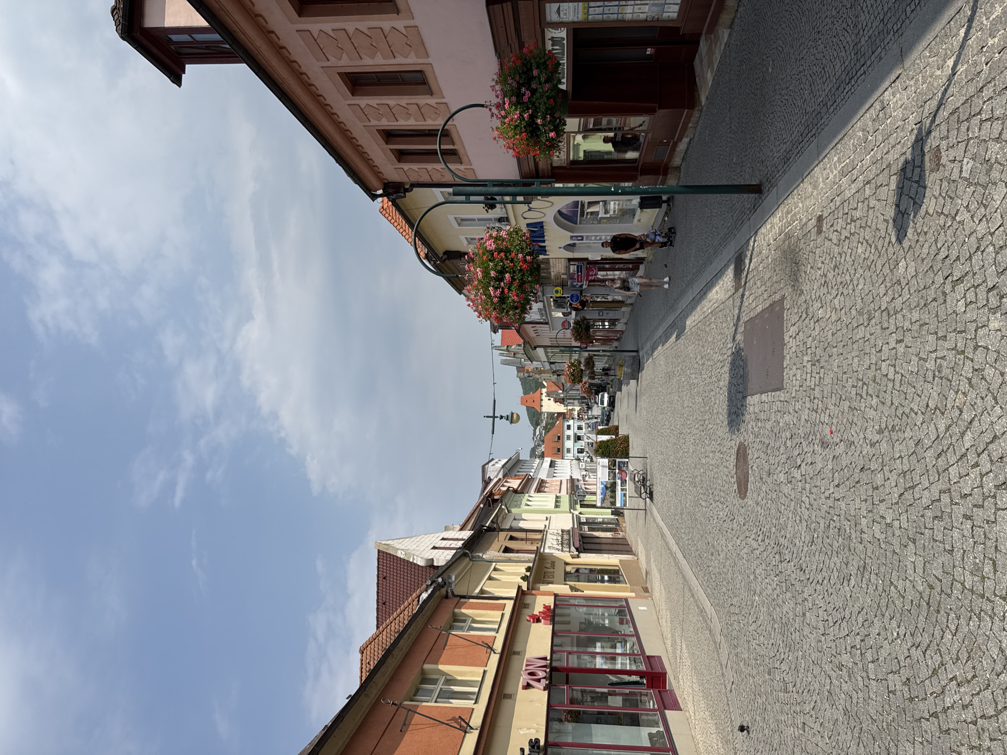 The image depicts a street scene in Beroun, Czechia, likely in the historic town center. The perspective is from a slightly elevated viewpoint, looking down a cobblestone street lined with buildings on both sides. The buildings are a mix of architectural styles, with some featuring colorful facades and red-tiled roofs. The street is relatively narrow and appears to be pedestrian-friendly, with no cars visible on the main thoroughfare.
In the foreground, the cobblestone pavement is the dominant feature, with a pattern of curved lines formed by the arrangement of the stones. A small black object, possibly a bird, is visible on the left side of the street. Further down the street, on the right side, a lamppost with hanging flower baskets is prominent. A couple, a woman and a man, are walking towards the viewer, with the woman pushing a stroller. The man is carrying a small child.
On the left side of the street, there are storefronts with large windows. One storefront has signage that reads "ZON" in large red letters. On the right side, more storefronts are visible, with awnings and signage. In the distance, the street curves slightly, and more buildings can be seen, including what appears to be a tower in the far background. The sky above is partly cloudy, with a mix of blue sky and white clouds. The overall impression is of a charming European town on a pleasant day.