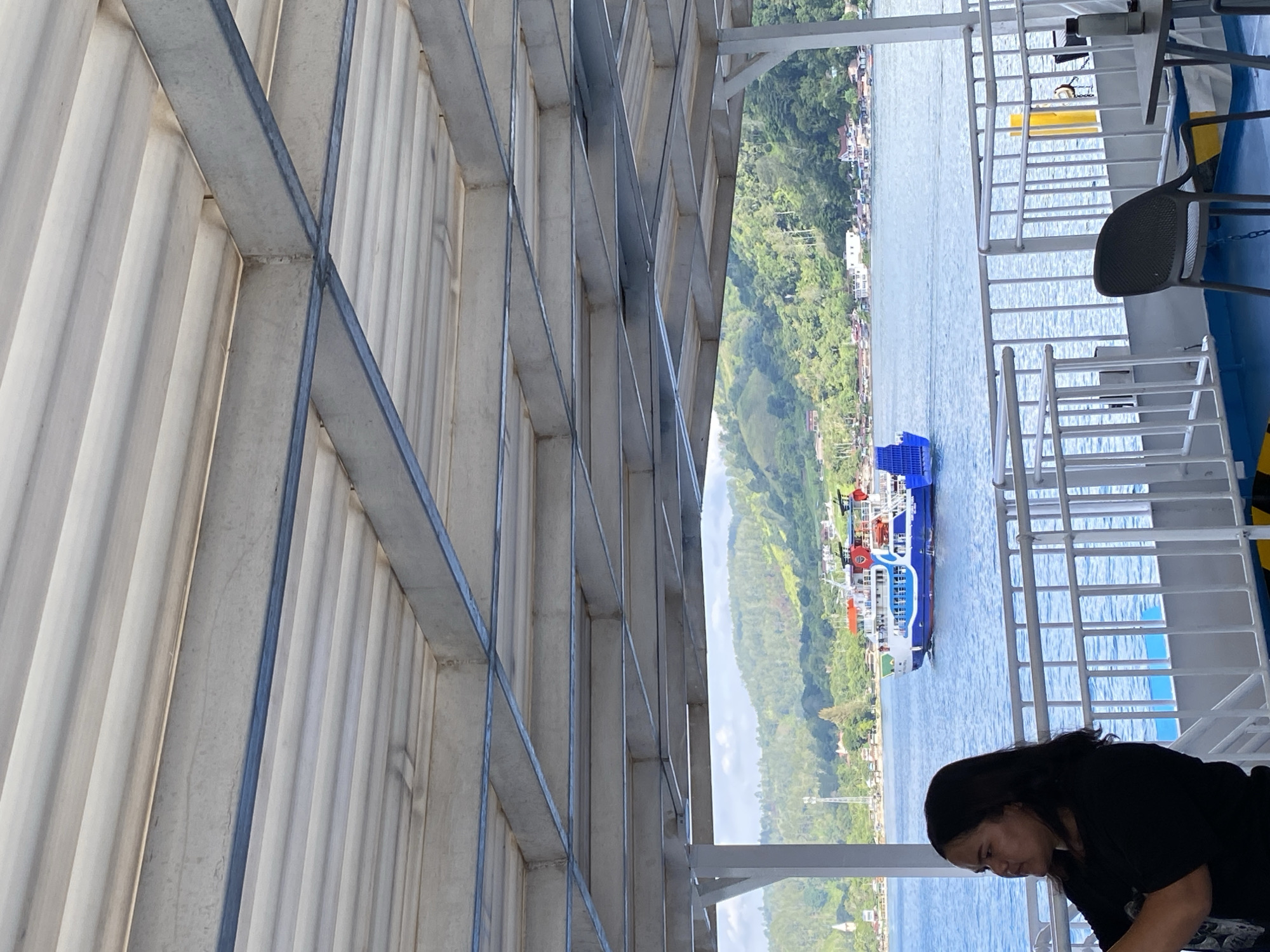 A woman with dark hair pulled back is standing on a deck, looking to the left with her arms crossed. She is wearing a black t-shirt. Behind her, a ferry with a blue hull and white and red upper decks is moving across a body of water. The water is calm and reflects the blue sky. In the background, a lush green hillside dotted with houses and trees slopes down to the water. The scene appears to be in the city of Parapat, Indonesia, likely on Lake Toba. The foreground of the image is dominated by the overhead structure of the ferry deck, with its metal beams and corrugated ceiling. A black plastic chair and a table are also visible on the deck.
