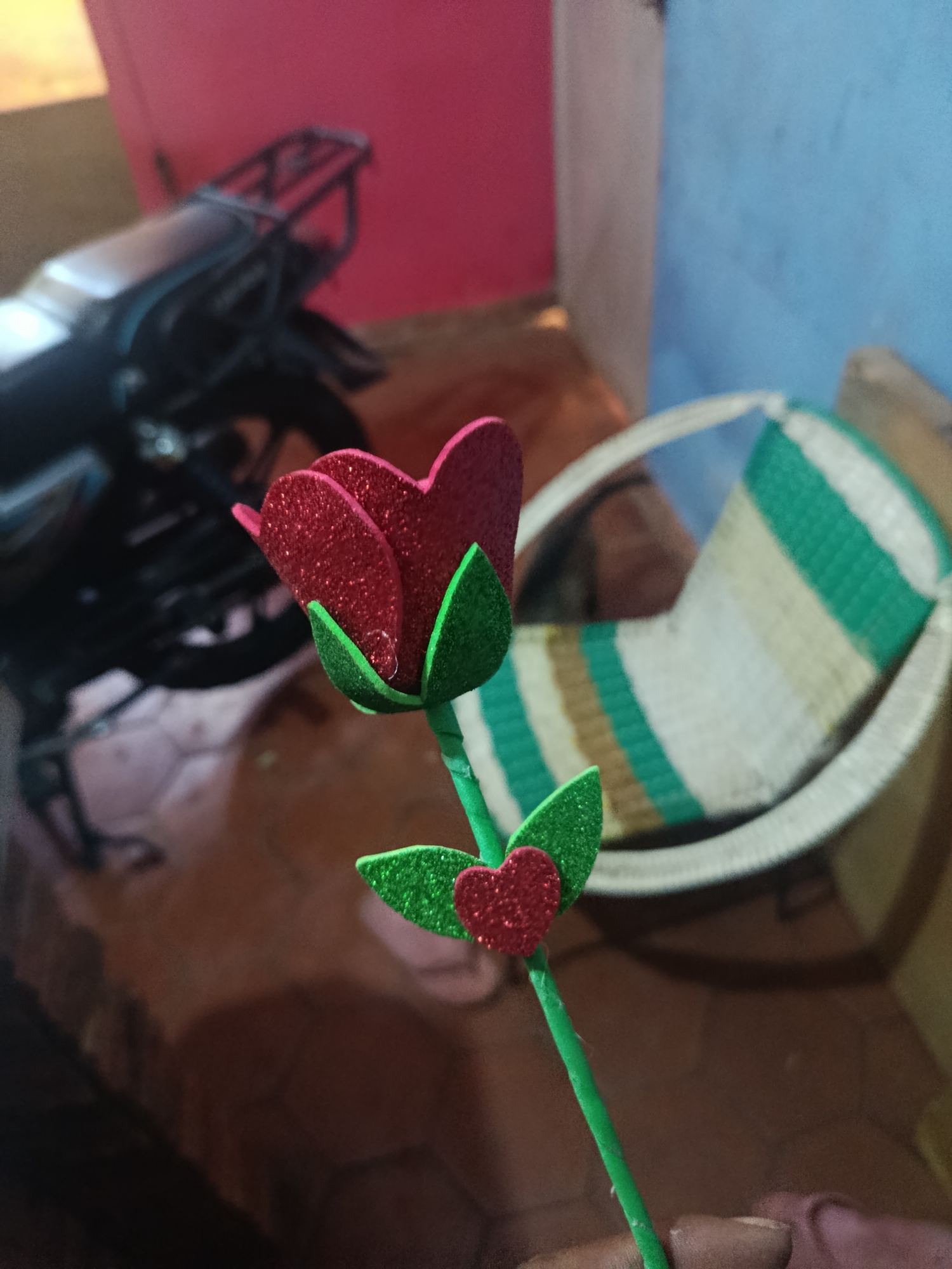 A close-up captures an artificial rose with red glitter-covered petals, green glitter-covered leaves, and a green stem. A glittery red heart adorns the stem just below the leaves. Behind the rose, partially blurred, is a motorcycle, a striped woven chair, and walls painted pink and blue. The scene appears to be indoors, possibly in a home. There is no way to confirm the location is Juan Griego, Venezuela, from the given image.