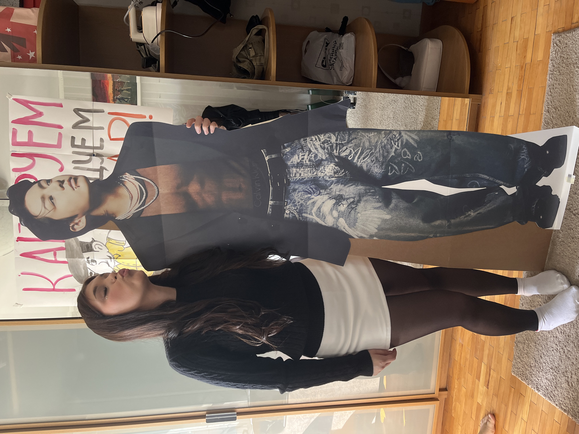 The image shows a young woman playfully interacting with a life-sized cardboard cutout of a male figure in an indoor setting, likely in Saint Petersburg, Russia. The woman, with long dark hair, wears a black long-sleeved top, a white skirt, dark tights, and white ankle socks. She poses with pursed lips, looking at the cutout, with her hands placed on its side.
The cardboard cutout depicts a male, possibly a celebrity or K-pop idol, wearing a dark blazer over a bare torso, exposing pearl-like necklaces. He has jeans with printed graphics, and the "Calvin Klein" logo is visible on his waistband.
The background features a wooden shelving unit holding various items, including a white plastic bag with the Russian text "СЕМЕЙНЫЙ МАГАЗИН" (Family Store) and a small appliance. Behind the cutout, a white banner or poster on the wall displays large, handwritten Russian text: "РАДУЕМ ЧЕМ Д!". The room has wooden flooring and a light-colored rug. Partial glass-fronted cabinets are visible in the lower left, and a bare foot is seen in the bottom right. The lighting indicates it is daytime, and the overall atmosphere is lighthearted and expressive.