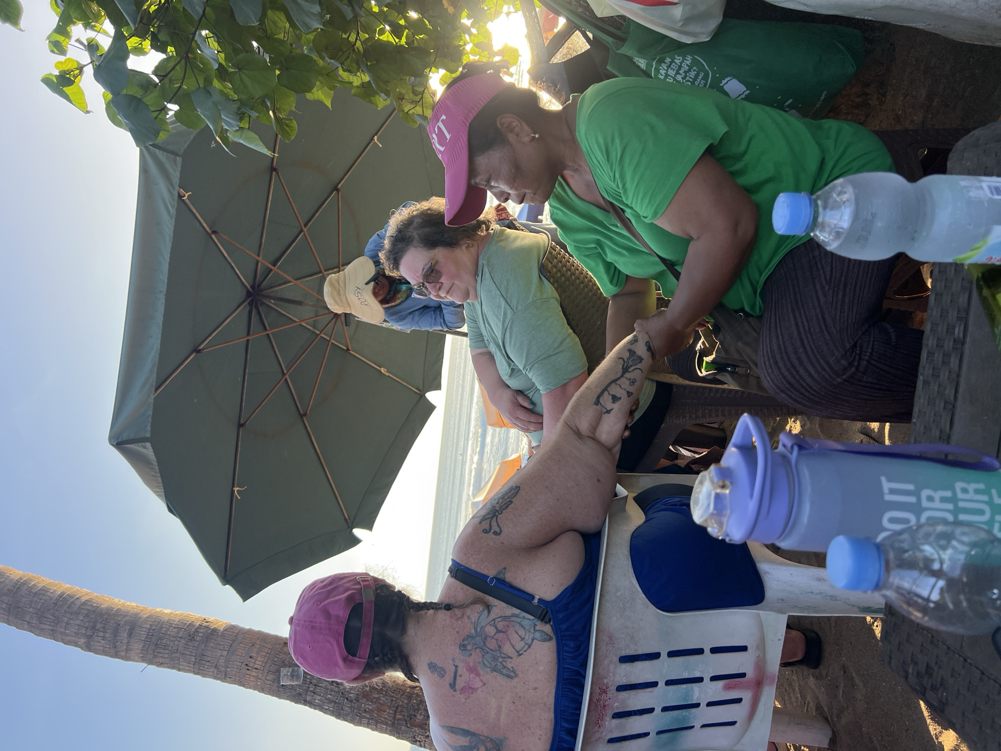 Four individuals are gathered outdoors, seated on chairs under a large, green umbrella. The setting appears to be a beach with sand visible on the ground, and the ocean and sky are in the background. The sun is setting, casting a warm glow and lens flare over the scene.
In the foreground, to the left, a person wearing a pink baseball cap and a blue tank top is seated, facing away from the camera. Their back is adorned with multiple tattoos, including a prominent turtle design. Next to them, a purple water bottle with gradient colors and a blue-capped clear plastic water bottle are placed on a dark-colored surface.
In the middle ground, two women are seated. The woman on the left, wearing a light green t-shirt and sunglasses, is looking upwards and to the left. The woman on the right, wearing a bright green t-shirt and a pink visor, is looking to the right. Their hands are clasped together.
In the background, another person, wearing a hat and a blue shirt, is visible standing behind the umbrella. They are looking down.
A palm tree trunk is present on the far left of the image, and foliage from a tree is visible on the right, partially obscuring the background. Two bags, one green with text, are placed near the woman in the green shirt. The overall atmosphere suggests a relaxed gathering during sunset.