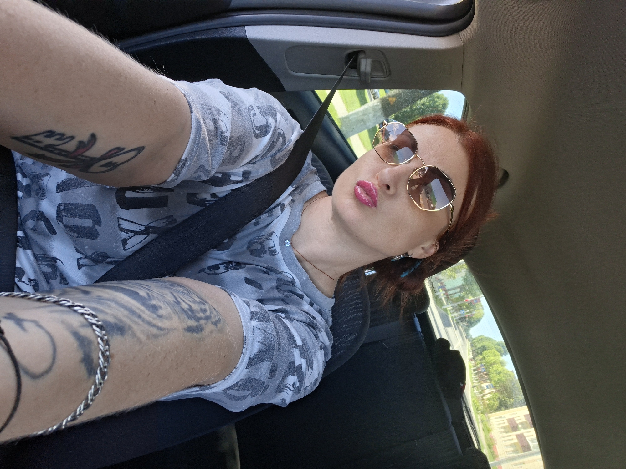 In a passenger seat of a car, a woman with red hair, wearing sunglasses and a light-colored, patterned shirt, puckers her lips for a selfie. Her left arm, adorned with a tattoo, is extended towards the camera. A black seatbelt is fastened across her chest. Through the car window, a tree-lined street with buildings and a blue sky can be seen, indicating a location in the city of Myadzyel, Belarus. The interior of the car shows the roof lining and the back of a seat.