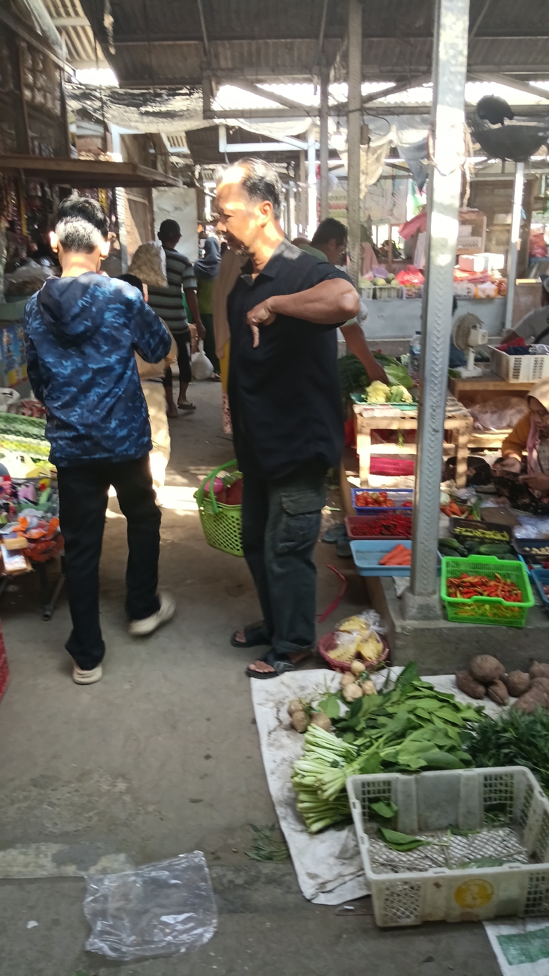 The image depicts a bustling open-air market scene, likely in Purwokerto, Indonesia. In the foreground, a man in a black polo shirt and cargo pants gestures with his right hand while facing a younger person in a blue patterned hooded jacket and black pants. Both are standing on a concrete floor. Behind them, various market stalls are visible, displaying an assortment of fresh produce including chili peppers, tomatoes, leafy greens, and root vegetables. Baskets and crates are used to hold and display the goods. Several other individuals can be seen in the background, engaged in shopping or working at the market. The environment suggests a typical Indonesian marketplace, with temporary structures and goods laid out for sale under a covered roof. Sunlight streams in from above, illuminating the scene.