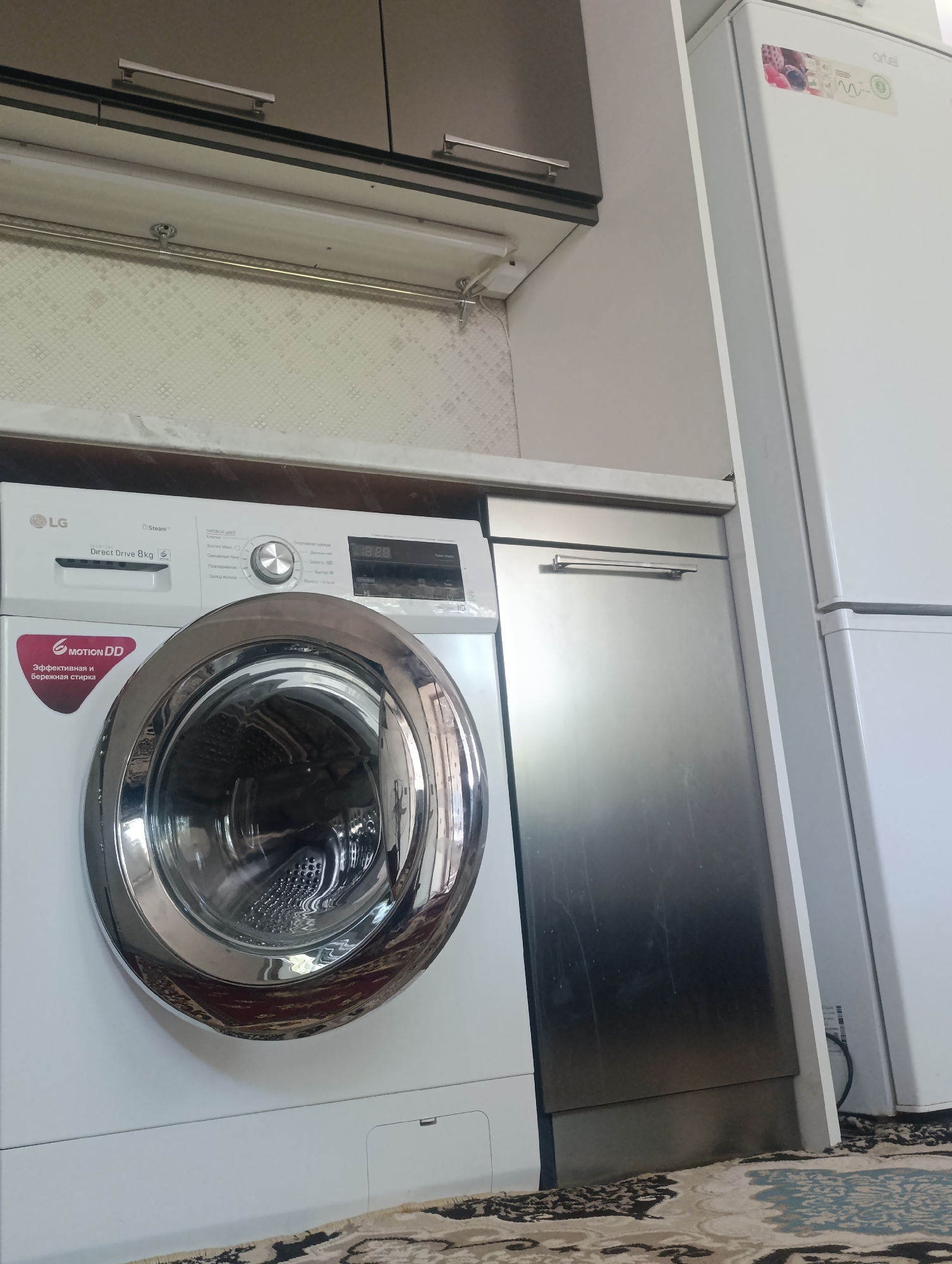 The image depicts a domestic laundry area or kitchen, likely within an apartment in Sovzharoq, Uzbekistan. Prominently featured is a white LG washing machine with a silver drum, positioned next to a silver-fronted appliance, possibly a dishwasher. To the right, a white refrigerator with some branding, including the "artel" logo, is visible. Above the appliances, a section of kitchen cabinetry with silver handles is present, along with overhead lighting. The background shows a textured, light-colored wall. The floor is covered by a patterned carpet. There are no people visible in the image, and no actions are taking place. The scene is static and appears to be a well-lit interior.