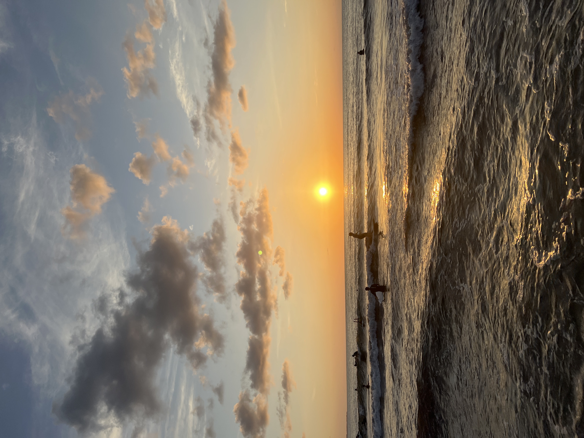 The image depicts a sunset over the sea in Isola delle Femmine, Italy. The sun, a bright orb, is positioned low in the sky, casting a warm, golden glow across the water and illuminating the clouds. The sky transitions from a pale blue at the top to a deep orange near the horizon. Several surfers are visible in the water, some paddling on their boards and others standing. The waves are gentle, with white foam cresting on their surfaces, reflecting the sunlight. The foreground shows the texture of the water with its shimmering reflections. The overall scene conveys a peaceful and serene atmosphere of a day ending at the beach.