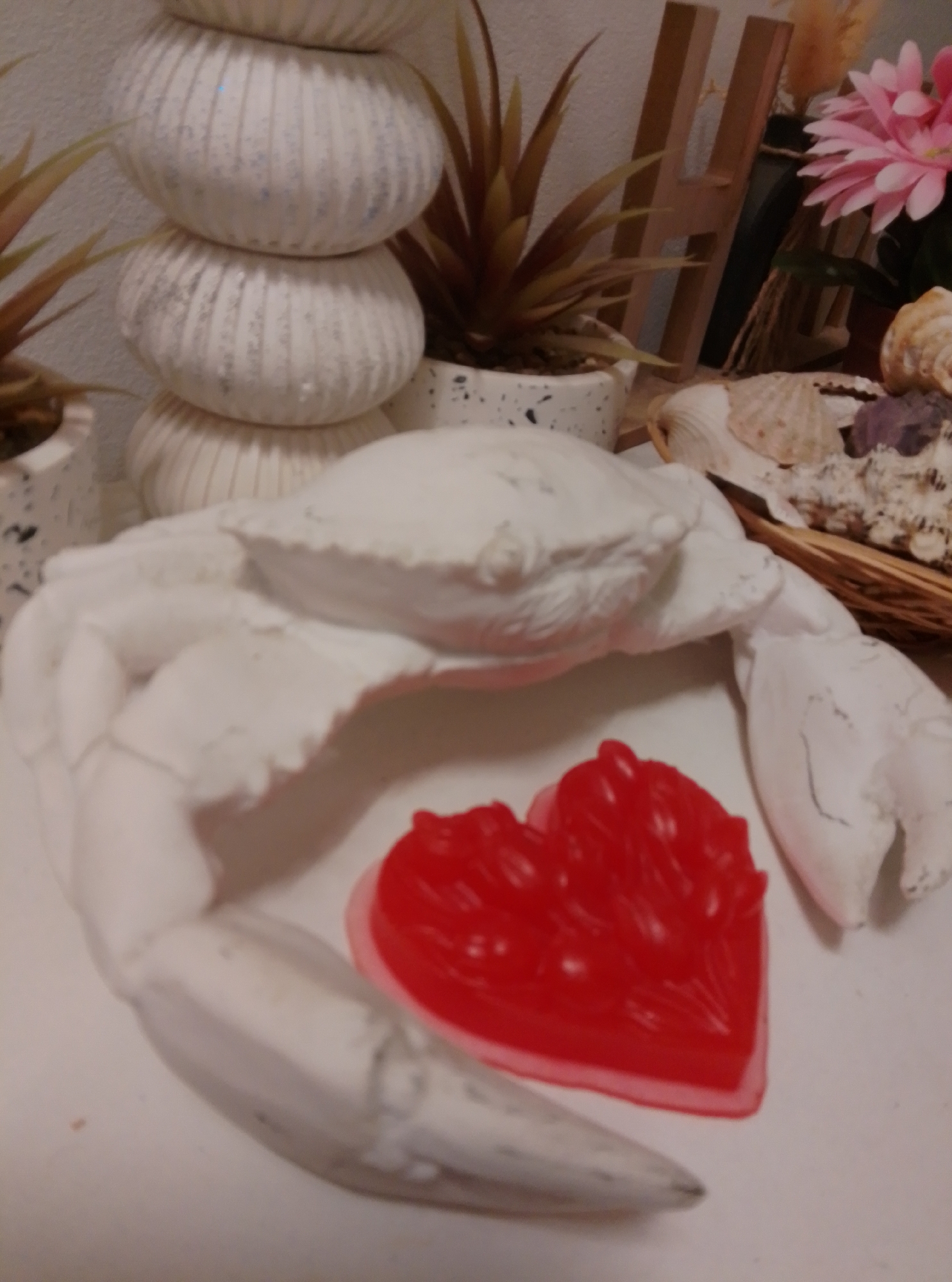 A white crab sculpture with a red heart-shaped soap is displayed on a white surface. The crab sculpture is in the foreground, with its claws extended. The red heart-shaped soap is placed in front of the crab. In the background, there are stacked white decorative pots, a wooden letter "H," some faux plants, and a basket filled with seashells. The scene is indoors and appears to be a home decoration. There are no people visible in the image. The image does not contain any information about the city of Wedel, Germany.