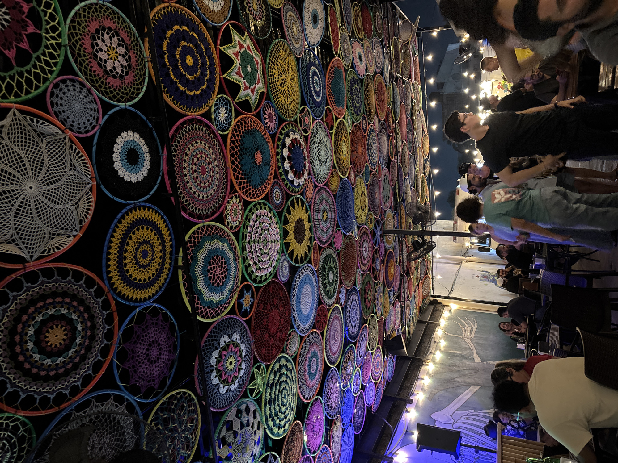 The image shows the interior of a bar/restaurant in Veracruz City, Mexico, at night. The left side of the frame is dominated by a wall covered in numerous colorful, circular crocheted or woven decorations, likely dreamcatchers or mandalas. A metal frame supports the decorative wall.
On the right side of the image, people are seen sitting at tables and standing, engaged in conversations. Overhead string lights are visible. The lighting is dim, with blue hues on the lower part of the frame. A mural featuring what appears to be a skeleton is painted on the wall behind the tables. A speaker is also visible in the lower portion of the frame.