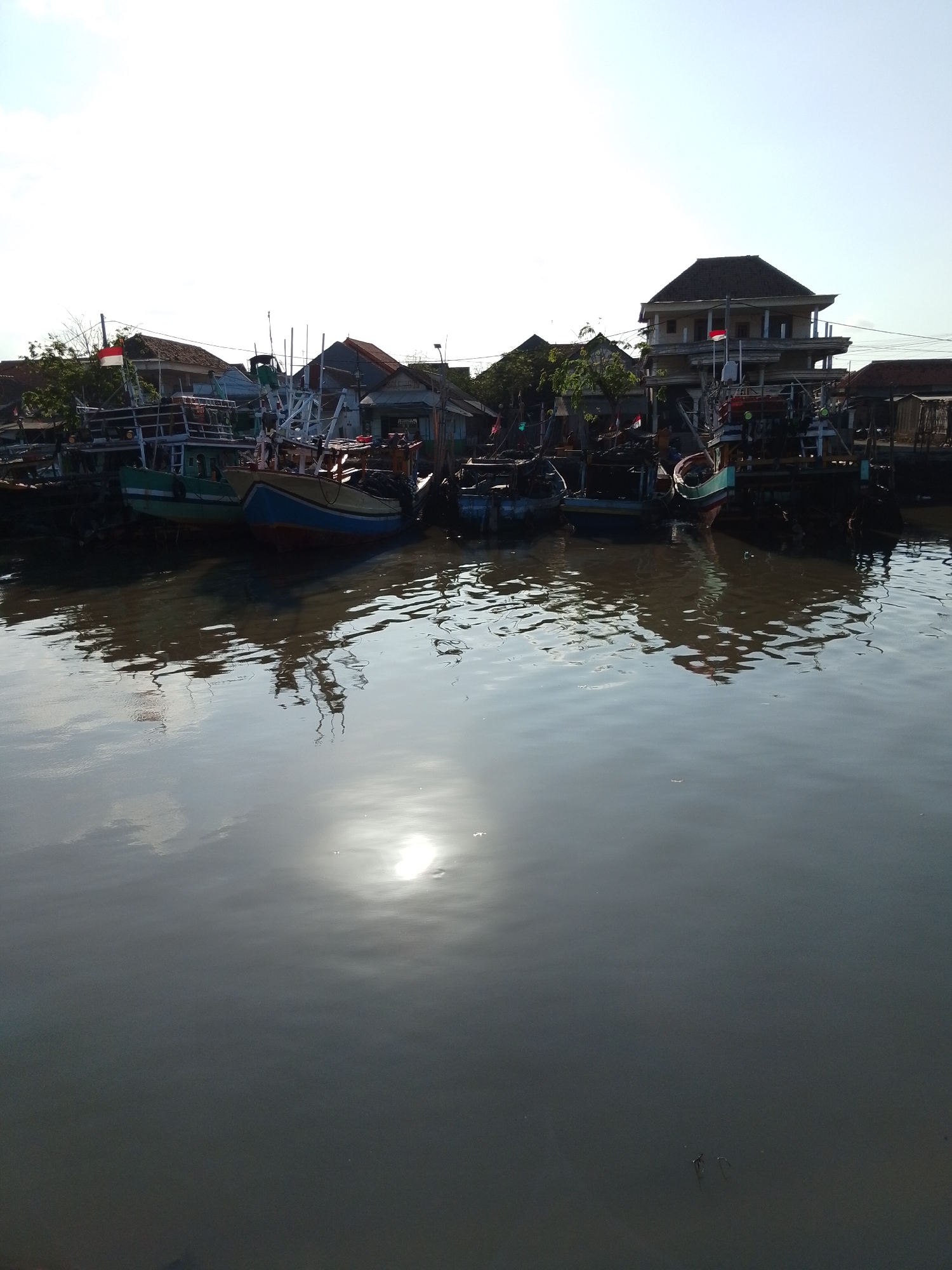 The image depicts a waterfront scene in Pasuruan, Indonesia. Several fishing boats are moored along the shore, with some appearing to be under repair or construction. A two-story building with a prominent dark roof stands behind the boats, flying the Indonesian flag. The water in the foreground reflects the boats, buildings, and the bright sky, with the sun creating a strong glare on the surface. The overall impression is of a working fishing community with some modern development.