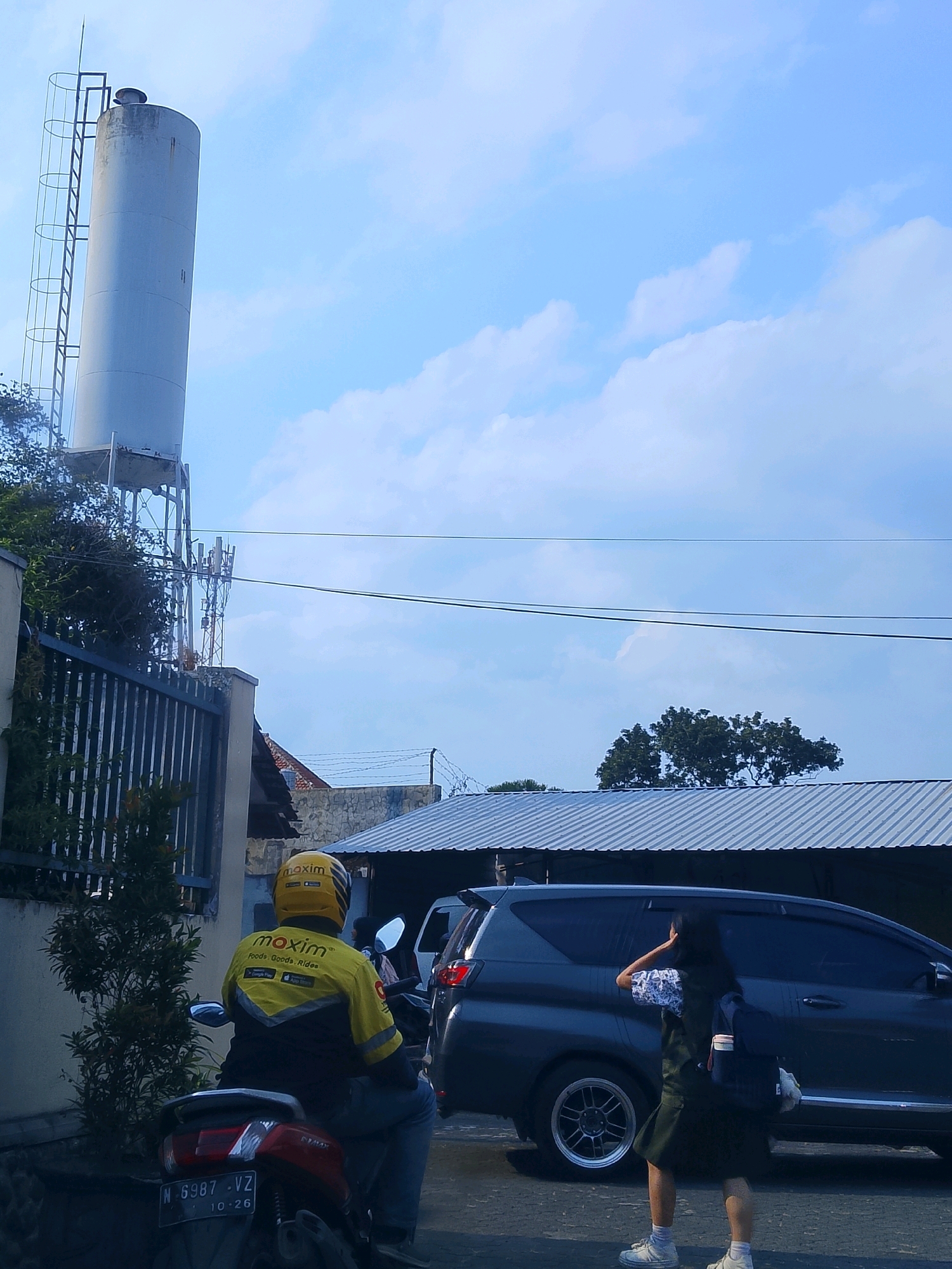 The image depicts a street scene in Malang, Indonesia, under a bright blue sky with scattered clouds. Dominating the upper left portion of the frame is a large, cylindrical water tower with a ladder attached to its side. To the right of the water tower, a cellular tower is visible. In the foreground, a person wearing a yellow jacket with "maxim" branding is seated on a red motorcycle. Behind the motorcycle is a dark-colored car. A young woman in a school uniform is walking towards the car, touching her hair. The environment suggests a residential or semi-urban area, with corrugated metal roofs visible on some buildings in the background. The license plate on the motorcycle is visible and reads "N 6987 VZ".