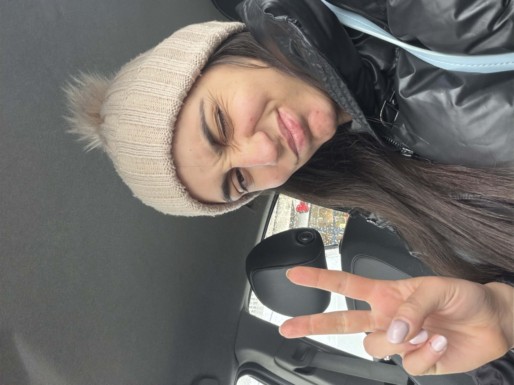A selfie shows a woman indoors, possibly in a car, making a peace sign with her right hand. She is wearing a light-colored knit hat with a pom-pom and a dark, puffy winter coat with a light blue strap. Her long brown hair falls over her shoulder. She is making a comical, squinting facial expression. Through the window, it appears to be a rainy or snowy day. Based on the location of the window frame, the car may be a Jeep Wrangler. Without more information, it is impossible to definitively determine the exact location within Sofia, Bulgaria.