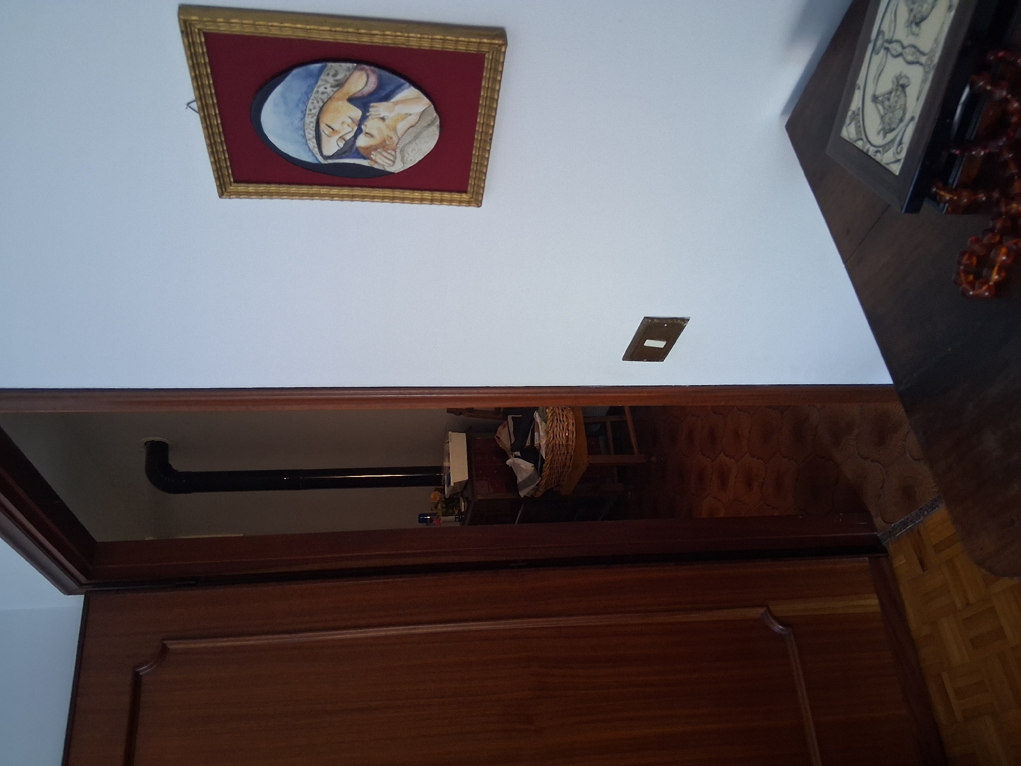 The image depicts an interior scene, likely a hallway or entryway, with a doorway on the left leading to another room. The doorway on the left reveals a section of a wooden door with decorative molding. The doorway to the right shows a glimpse into a dimly lit room containing a dark wood dresser, a wicker basket filled with laundry, and a chair. A black metal pipe, possibly from a stove or chimney, is visible vertically in this room.
On the wall to the right of the main frame, a framed religious artwork is displayed. The artwork features an oval depiction of the Virgin Mary and the infant Jesus, rendered in muted colors with the figures appearing to be kissing or embracing. The frame is gold, with a dark red matting. Below the artwork, a light switch is mounted on the white wall.
In the lower right corner of the image, a dark wood surface, possibly a table or desk, is partially visible. On this surface rests an open decorative box with an intricate patterned inlay. A string of amber-colored beads is draped across the box and onto the surface.
The lighting in the scene is moderate, with the room on the right appearing darker than the foreground area. The floor in the foreground appears to be parquet, while the floor in the room on the right has a patterned tile design. The overall impression is one of a lived-in, traditional home environment. The image appears to be a photograph taken from within the city of Colorno, Italy.