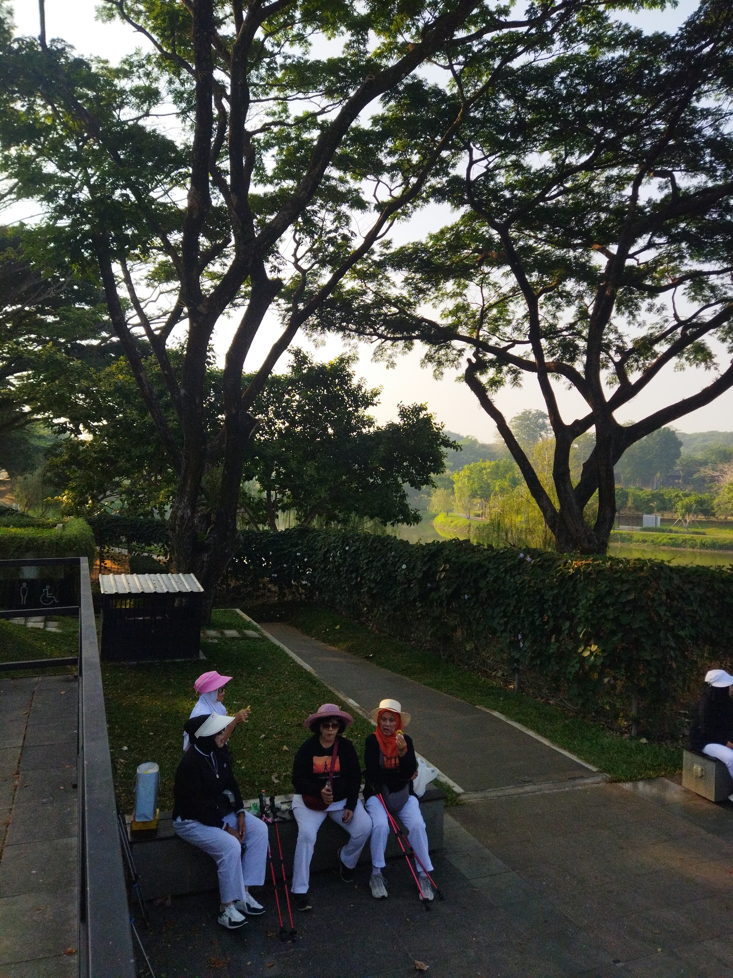 The image depicts a group of four women dressed in athletic wear and hats, some with headscarves, taking a break on a bench in a park setting. They appear to be engaged in or preparing for an outdoor activity, as two of them are holding walking poles. The background features lush greenery, tall trees, and a body of water, suggesting a scenic park or botanical garden. The overall atmosphere is relaxed and casual. Based on the context provided, this scene is located in a park within the city of Semarang, Indonesia.