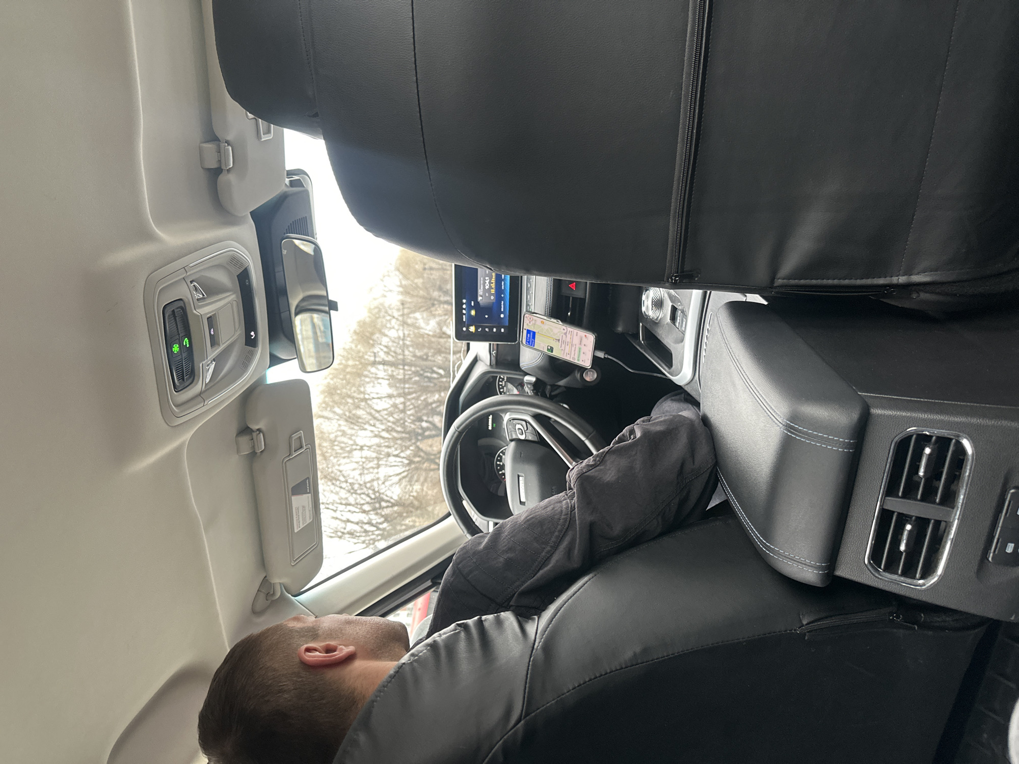 A man is reclining in the driver's seat of a vehicle. He appears to be resting or sleeping. The interior of the vehicle is visible, including the dashboard, steering wheel, infotainment screen, and rear air vents. A smartphone is mounted on the dashboard, displaying a navigation app. The location is not discernable.