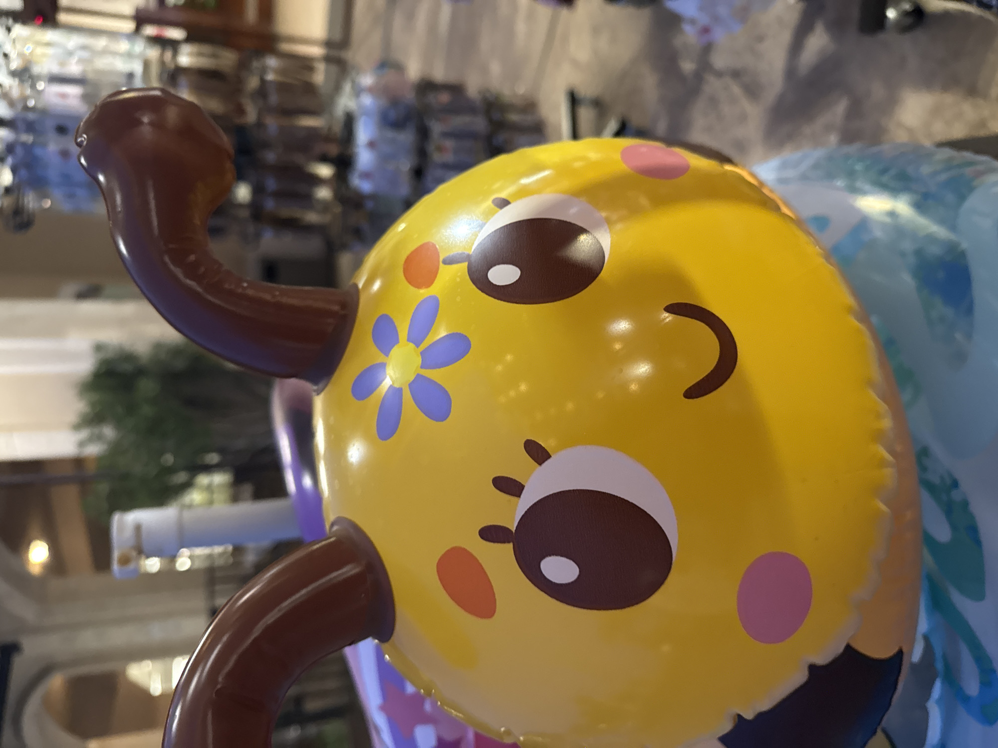 A close-up, angled shot captures a large, yellow inflatable character, resembling a stylized bee or insect, positioned in what appears to be an indoor or covered public space. The character features prominent, oval-shaped cartoon eyes with white highlights, giving it a cheerful and friendly expression. A simple curved line forms a smiling mouth.
Decorating its round, shiny yellow surface are several colorful decals: a purple flower with a yellow center near one eye, a light orange blush mark on one cheek, and a pink blush mark on the other. Two glossy, brown, curved antenna-like appendages extend from its head, one prominently displayed on the left side of the frame. The inflatable's surface shows minor texture and subtle reflections, indicating its material.
In the blurred background, hints of architectural elements like arches or pillars, indistinct light sources, and possibly some greenery are visible, suggesting a well-lit environment such as a mall atrium, a large hotel lobby, or a similar spacious interior. A portion of a blue and white object, possibly another part of the inflatable or a separate decoration, is faintly visible behind the main subject. No text is discernible in the image.
