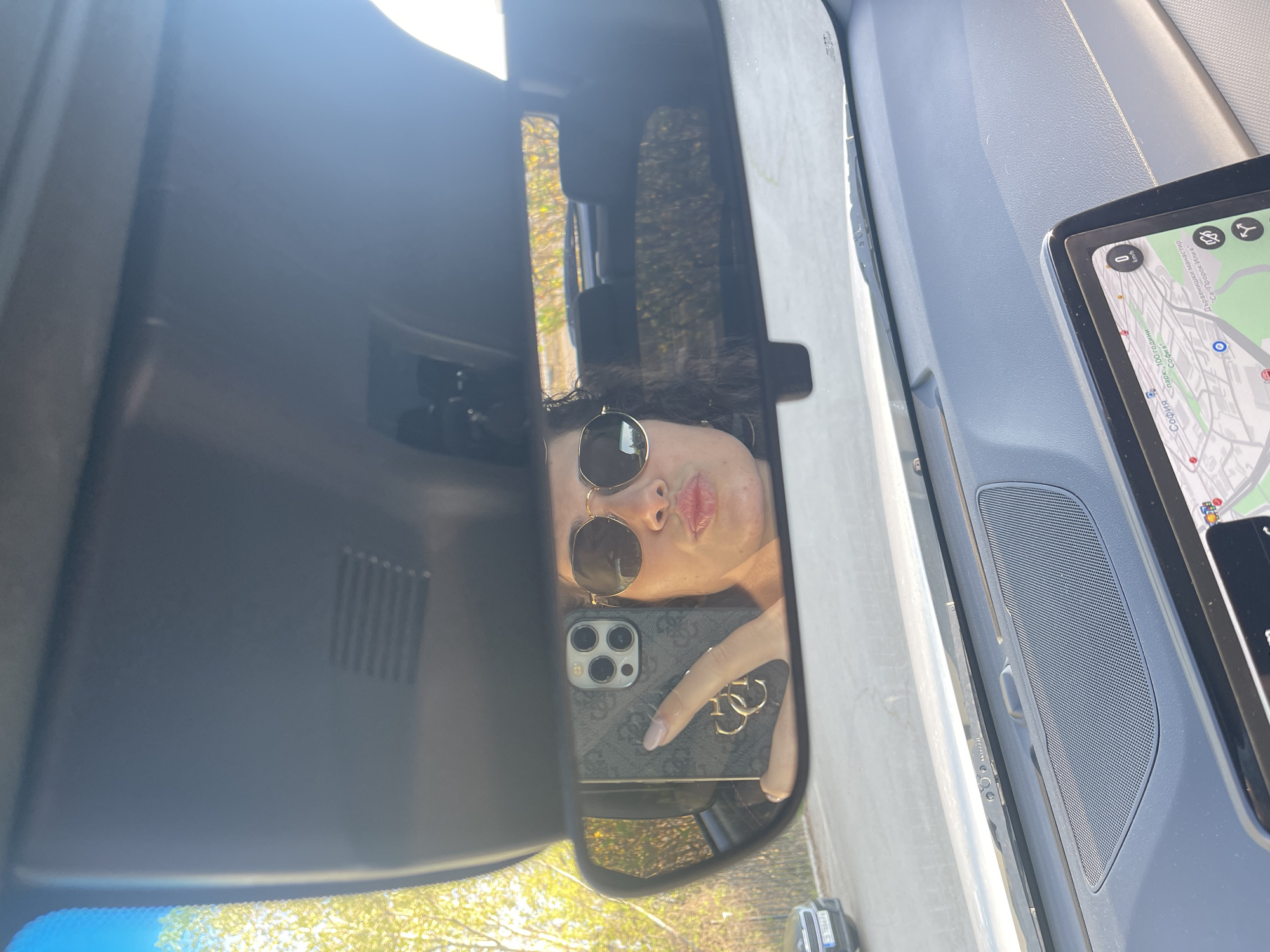 A mirror selfie is captured inside a car during daytime. The image is oriented vertically, centered on the car's rearview mirror. Reflected in the mirror is a person, likely female, with dark, curly hair, wearing round sunglasses and making a playful, puckered lip expression. She is holding an iPhone with a multi-lens camera setup, housed in a dark "Guess" branded case featuring a prominent logo and gold ring holder. Her fingers, adorned with light nail polish, grip the phone.
The background reflected in the mirror shows bright sunlight filtering through trees, indicating outdoor, sunny conditions. To the right of the rearview mirror, a car dashboard is visible, featuring a large tablet or navigation screen. The screen displays a map with Cyrillic text, clearly showing "София" (Sofia) multiple times, along with landmarks like "парк, 100 години" (Park, 100 years), "Светогорски манастир" (Svetogorski monastery), and "Пророк Илия" (Prophet Elijah), confirming the location as Sofia, Bulgaria. A speed indicator on the map shows "0 km/h". The car's interior appears modern with dark grey plastic and a textured speaker grille visible on the passenger side dashboard.