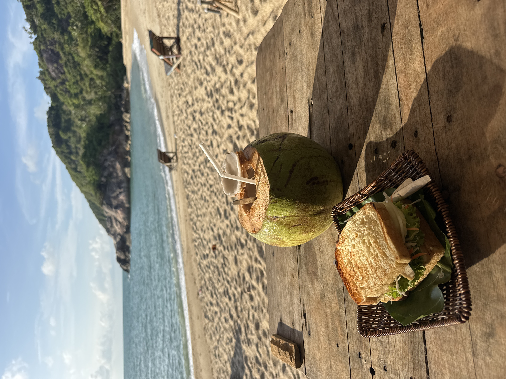 A photograph captures a still life scene set against the backdrop of a beach in Baan Thong Nang, Thailand.
The foreground features a wooden deck on which a green coconut, pierced with a straw, and a sandwich presented in a wicker basket are placed.
The background reveals a sandy beach that meets the sea, while a landform with lush vegetation extends into the frame on the left, establishing the location. The sky is visible above the landform. Two chairs are visible on the beach.