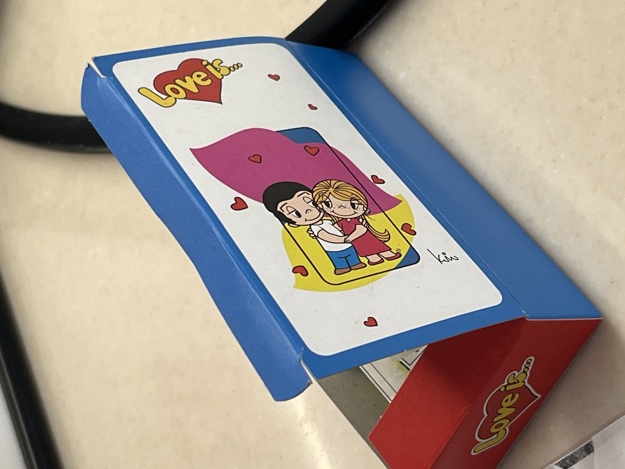 The image shows a "Love is..." gum box on a light-colored surface, possibly a table or countertop. The box is primarily blue and red, with a cartoon illustration of a couple embracing on the front, surrounded by hearts. The "Love is..." logo is prominently displayed on both the front and side of the box. A black cable or object is visible in the background. The photograph appears to have been taken indoors under artificial lighting. No information about the location in the city of Новое, Russia is apparent from the image.