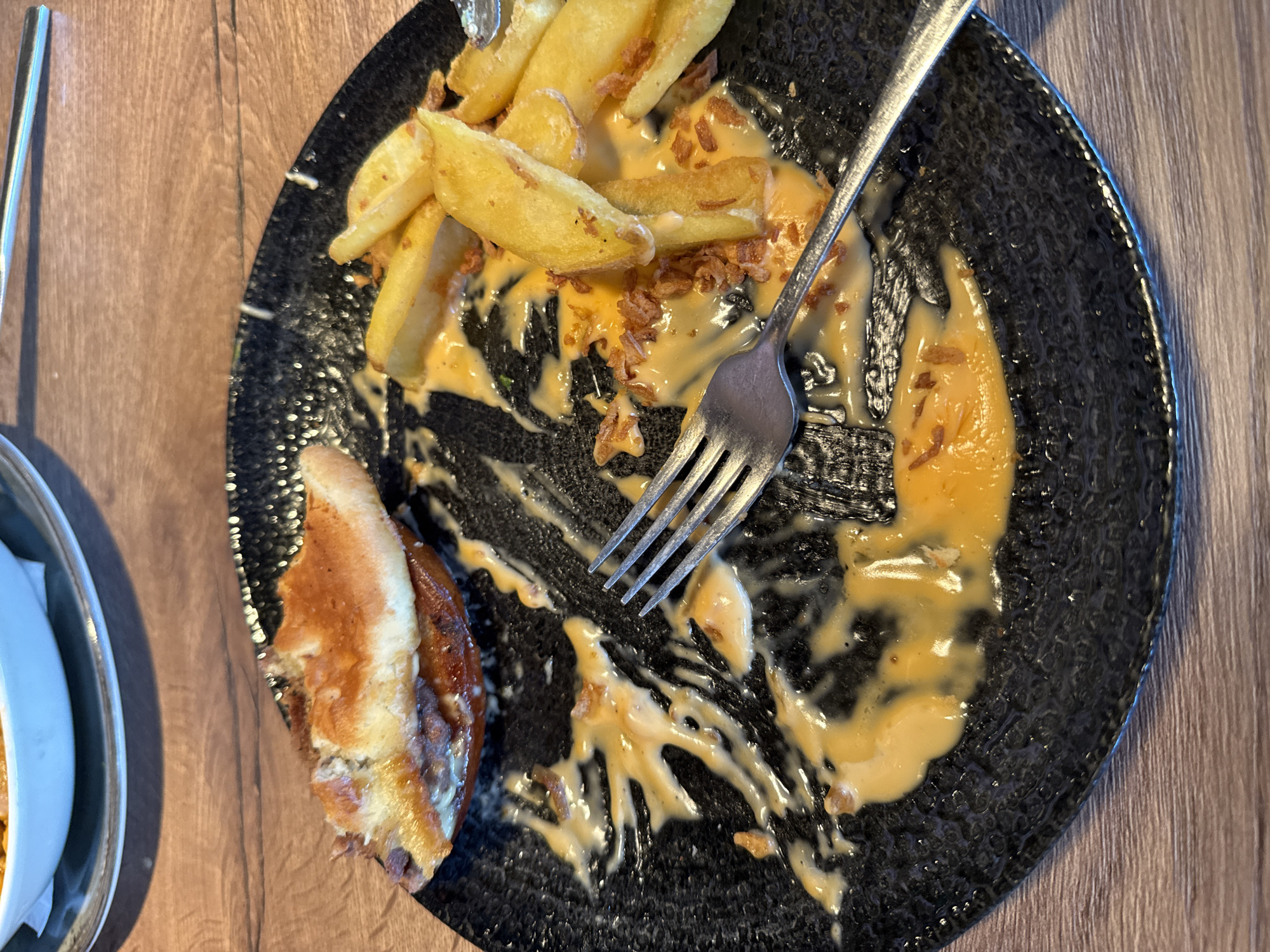 The image presents an overhead view of a partially eaten meal served on a dark, textured plate. The primary subjects are the food remnants, consisting of several chunky potato wedges generously coated in a creamy, light orange-yellow sauce, suggestive of a cheese sauce. Mixed with the sauce and fries are small, crispy, dark brown pieces, possibly fried onions or bacon bits. A silver fork, stained with some sauce, rests diagonally on the plate, indicating that the meal is nearing its conclusion.
On the left edge of the plate, a toasted portion of a bun or bread roll is visible, implying the meal likely included a burger or similar item. The dark plate has a subtle, raised, patterned surface. The meal is set on a wooden table, characterized by visible grain lines. A small section of another metallic or ceramic dish is partially visible on the far left.
The scene captures the aftermath of a casual dining experience. There is no visible text in the image. While the location context is Radinovo, Bulgaria, no specific visual cues within the image confirm this. The lighting suggests an indoor setting, but the specific time of day is not discernible.