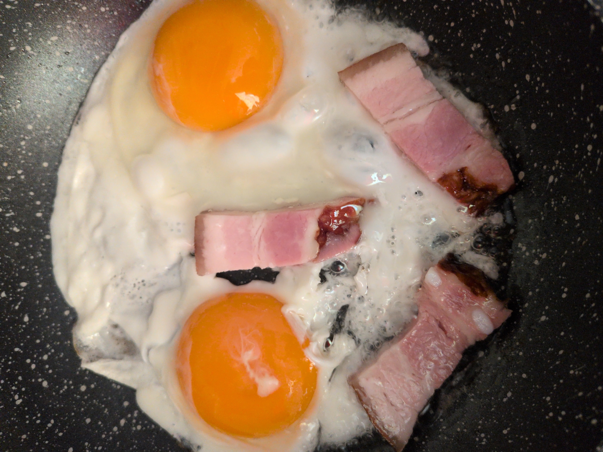A close-up, overhead shot shows two fried eggs with bright orange yolks and a foamy white base, cooking in a black speckled non-stick frying pan. Three strips of pink and white bacon are also cooking in the pan, arranged in a smiley-face configuration with the eggs acting as the eyes and the bacon strips forming the mouth. Bubbles are forming in the cooking egg white, indicating that the food is hot and being prepared. The background is the dark, speckled surface of the frying pan. The scene is domestic and pertains to food preparation. No specific location in Sochi, Russia, can be identified.