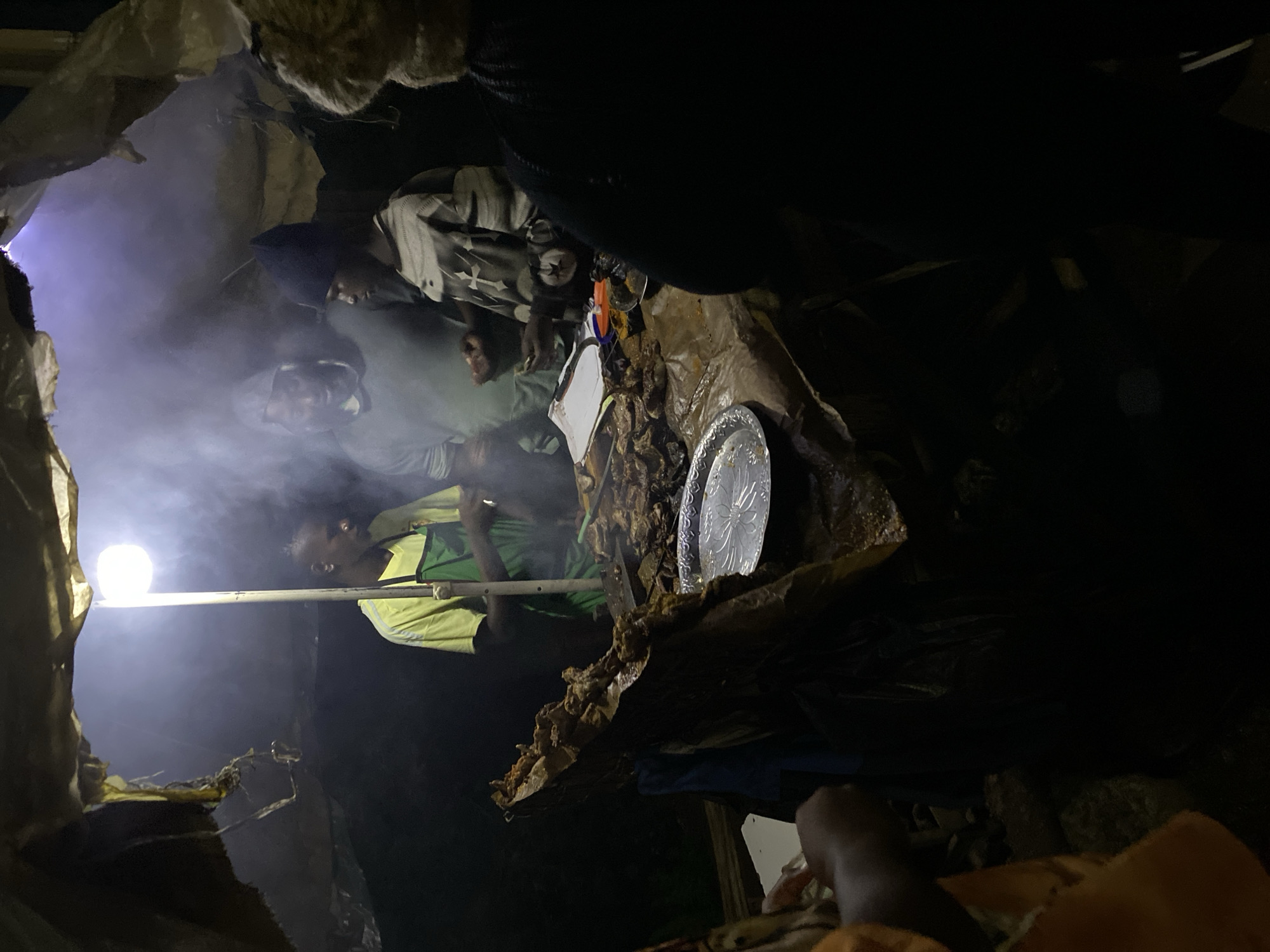 The image depicts a dimly lit street food stall in Bukuru, Nigeria, possibly at night given the artificial lighting. The stall is covered by a makeshift canopy, made of what appears to be tarp or corrugated metal, creating a somewhat enclosed space. A bright, round light fixture hangs from the center of the canopy, illuminating the scene and casting a strong light on a pile of meat and smoke.
Three individuals are visible behind a table laden with food. The person on the left, wearing a bright yellow t-shirt and a green apron, is facing towards the meat and appears to be preparing or serving it. To their right, a person in a grey hooded sweatshirt and a dark beanie is looking down at the table. Another person, wearing a patterned long-sleeved shirt and a dark beanie, is standing to the right of the person in the grey sweatshirt, also looking at the table and holding what might be money or food. A fourth person, partially visible on the far right and wearing a dark jacket and fur-lined hat, is facing away from the camera and towards the stall.
The table surface is covered with a plastic or paper sheet, and on it are several plates of grilled or roasted meat, some appearing to be large cuts. A metallic plate with a floral pattern sits prominently in the foreground on the table, holding a smaller portion of meat. There is also a stack of white paper or a notepad on the table, along with some utensils. Smoke is rising from the food and the cooking area, creating an atmospheric haze. The ground in front of the stall is dark and appears to be unpaved. The overall impression is of a busy, informal food preparation and sales environment.
